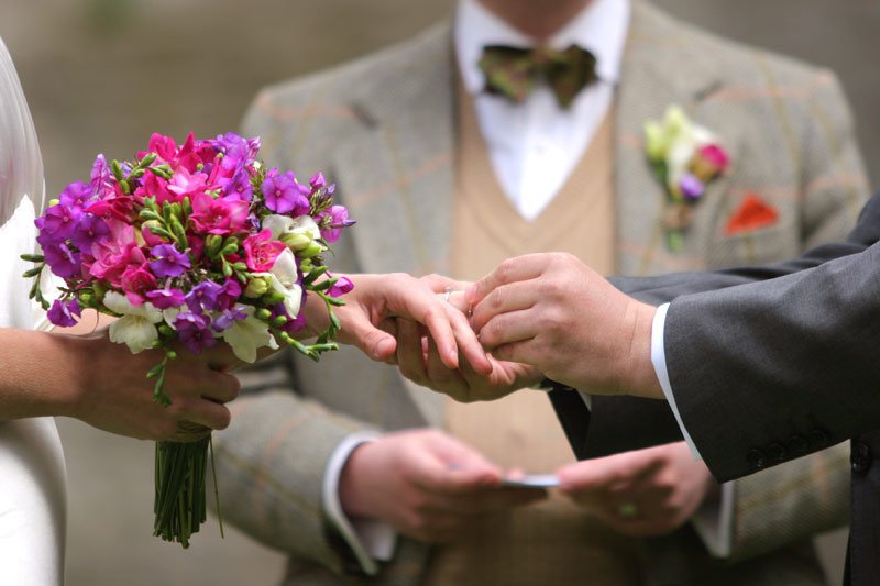 Close-up of a couple exchanging wedding rings during a ceremony, with a bridesmaid holding a bouquet of pink, purple, and white flowers in the foreground and a person in a suit in the background.