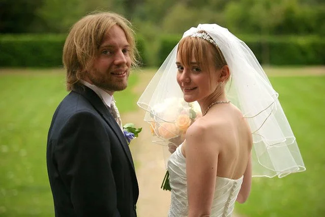 A bride and groom standing outdoors on a grassy area, smiling at the camera. The bride is wearing a wedding dress and veil, holding a bouquet of flowers. The groom is in a dark suit with a boutonniere.