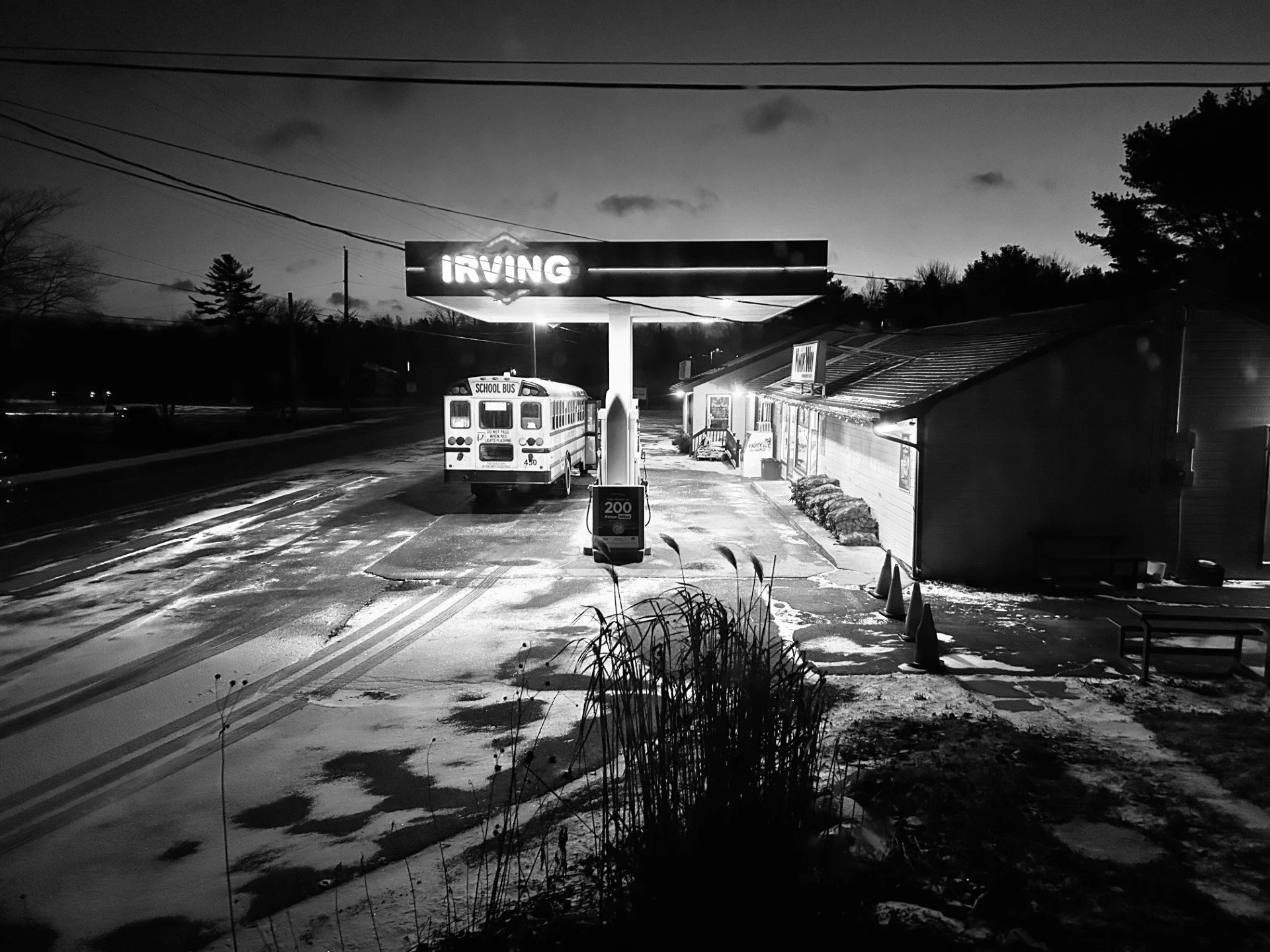 A black and white photo of a small town bus stop at night, with a school bus parked nearby, a lit sign reading 'IRVING,' a small building by the bus stop, and snowy ground.