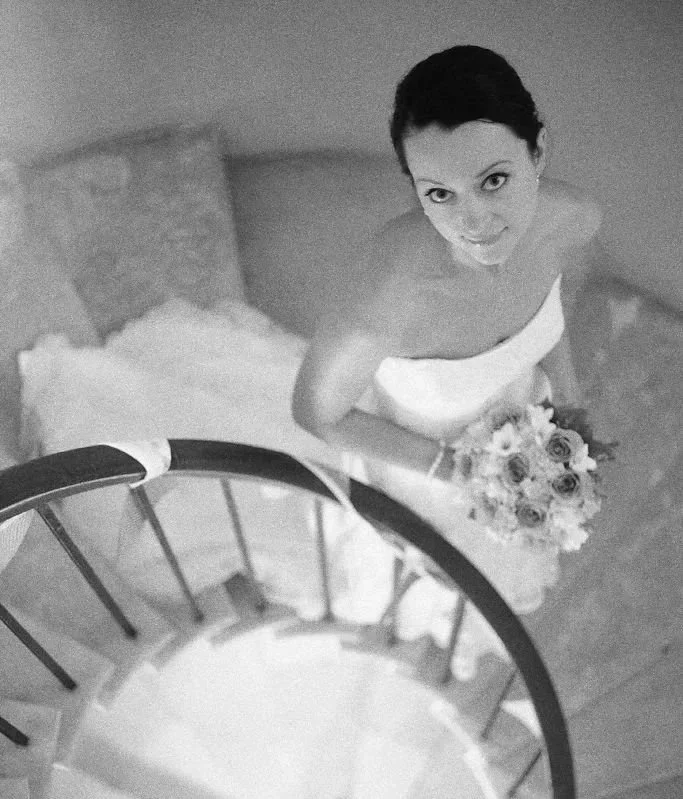 A black and white photo of a bride holding a bouquet, looking up at the camera from the top of a staircase.