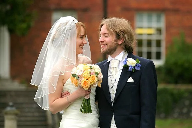 A bride and groom smiling at each other outdoors on their wedding day, with the bride holding a bouquet of yellow and white flowers and wearing a veil.