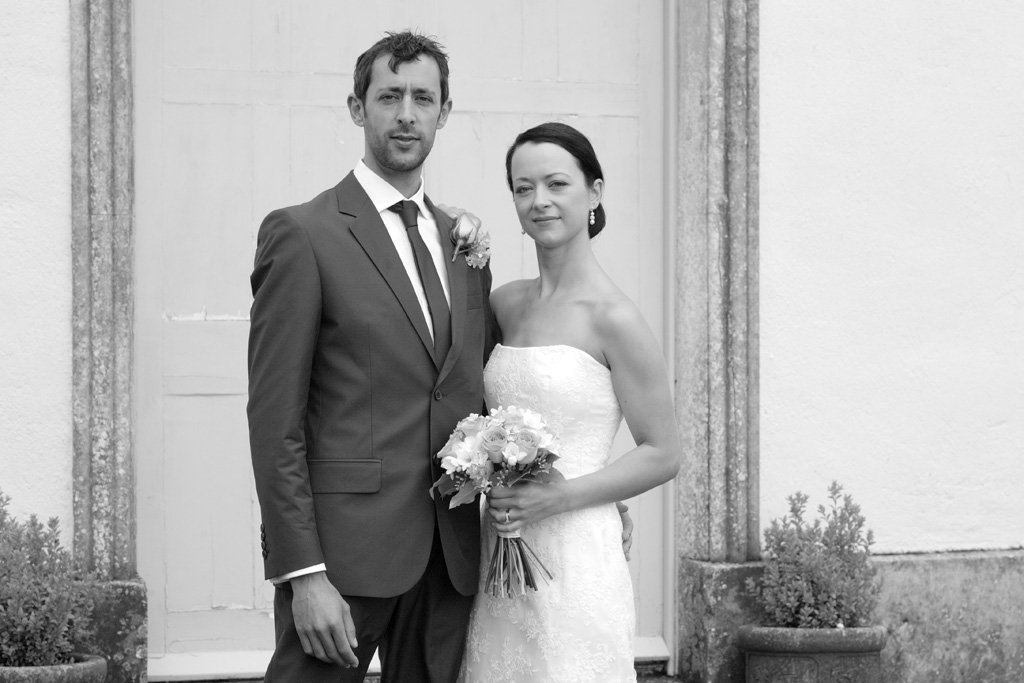 Black and white photo of a bride and groom standing close together, the groom in a suit and tie, the bride in a strapless wedding dress holding a bouquet of flowers, outdoors in front of a building.