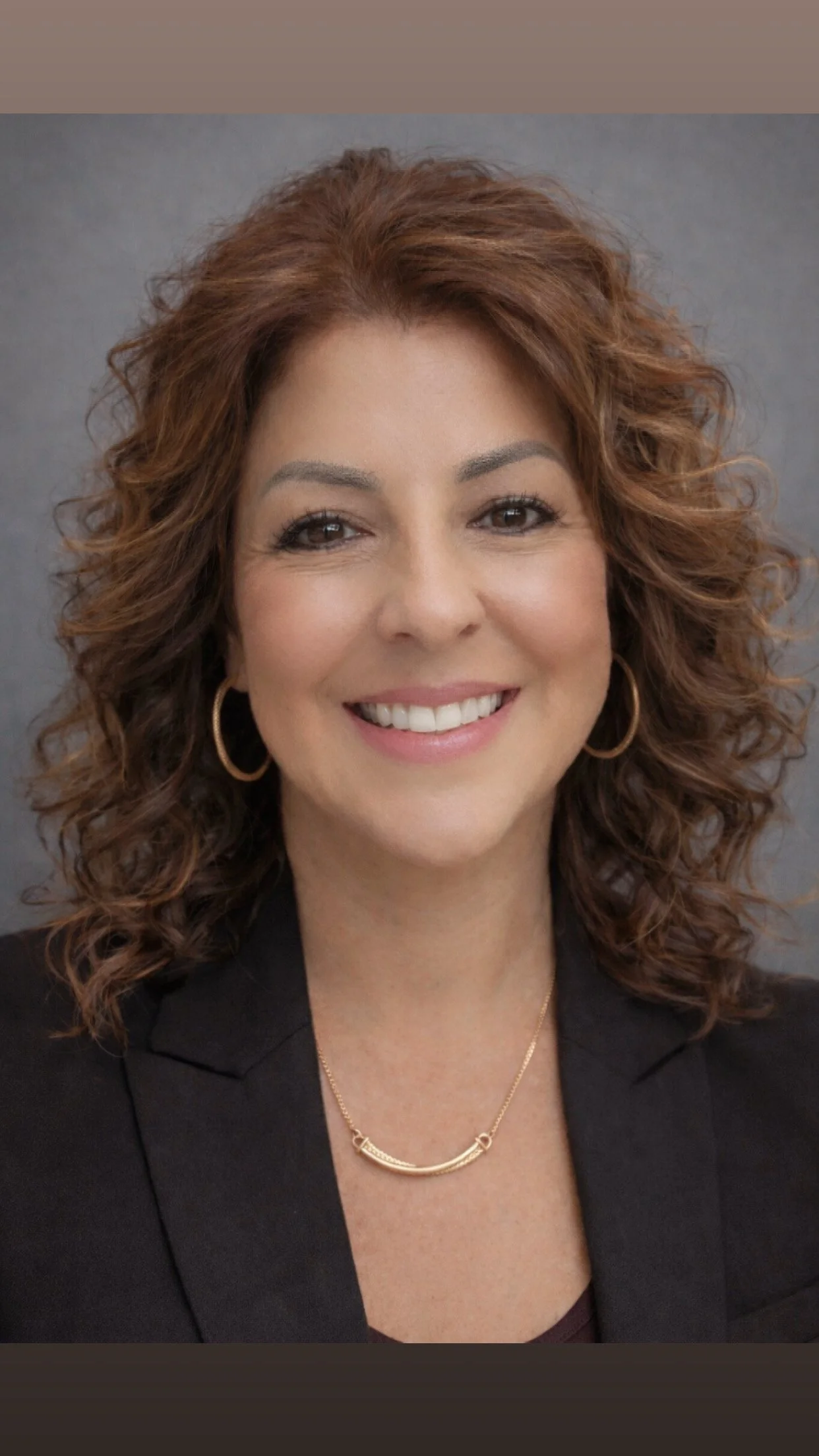 A portrait of a woman with curly, auburn hair smiling, wearing gold hoop earrings, a gold necklace, and a black blazer against a plain background.