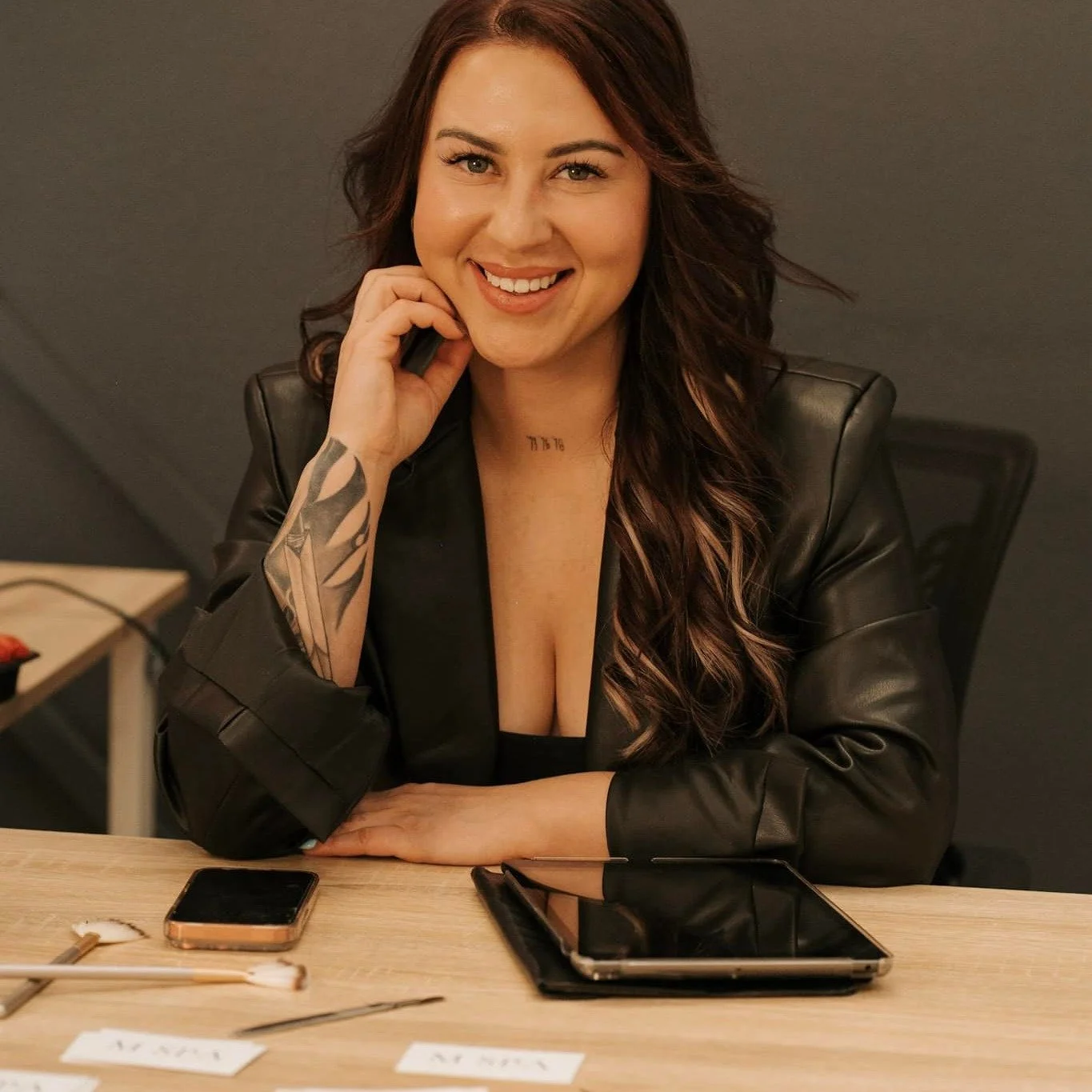 A woman with long, wavy hair, wearing a black leather jacket, sitting at a wooden table with a phone and tablet in front of her, smiling at the camera.