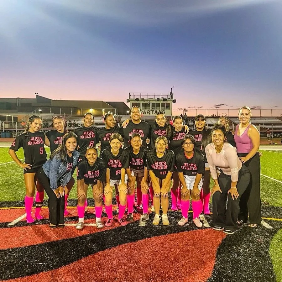 Youth girls' soccer team with coach and assistant on a football field during sunset, wearing black jerseys with pink accents and pink socks.