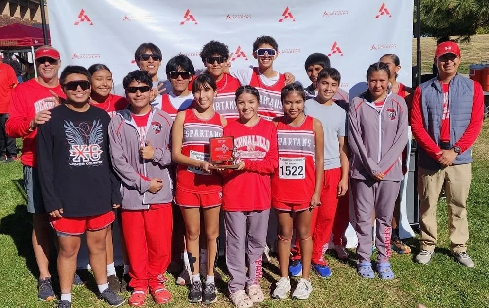 A group of young athletes and coaches in red and gray uniforms, standing outdoors on grass, posing for a photo after a race event. Some are holding a trophy, and there is a backdrop with logos.