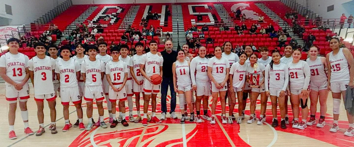 Girls high school basketball team in white uniforms with red accents standing on a basketball court, posing for a group photo with a coach and holding a trophy and a trophy box. Empty red bleachers and spectators are in the background.