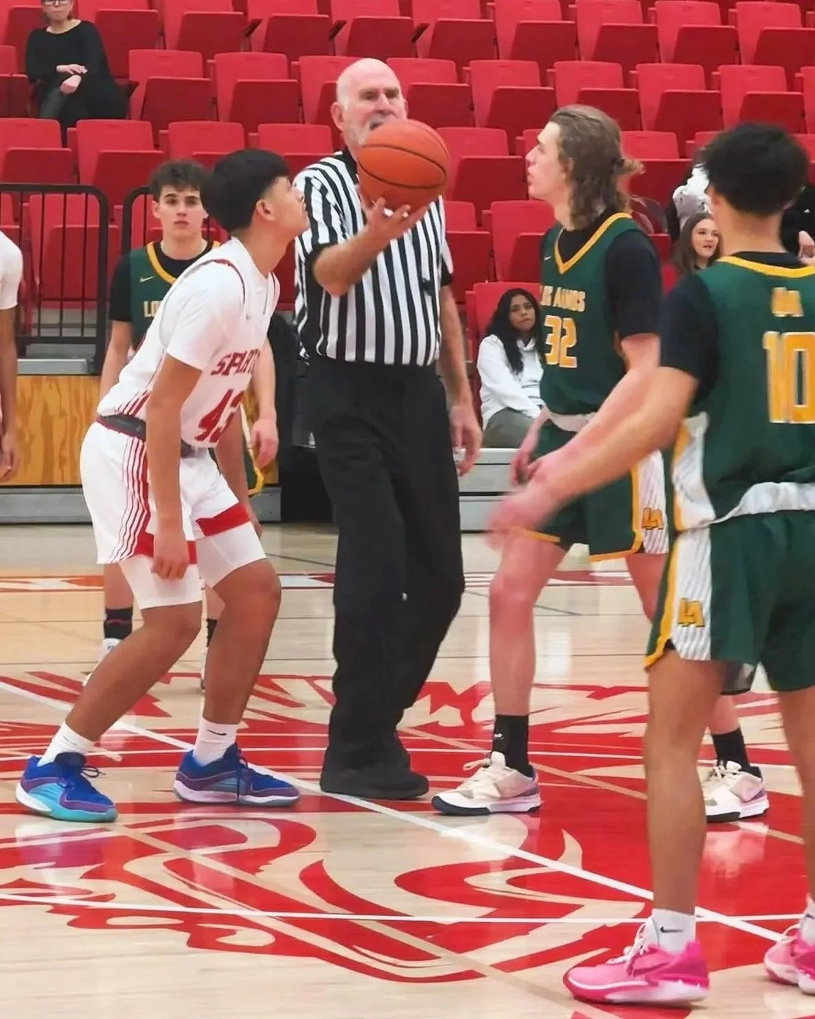 A basketball game coin toss at center court, with players in green and white uniforms, a referee holding the ball, and spectators in the background.