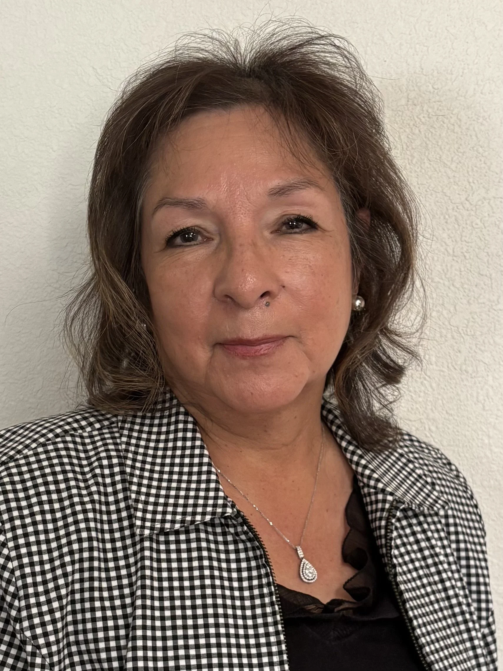 A woman with shoulder-length brown hair wearing a black and white checked blazer and a black top with ruffles, accessorized with a pearl earring and a teardrop-shaped pendant necklace, standing against a plain light-colored wall.
