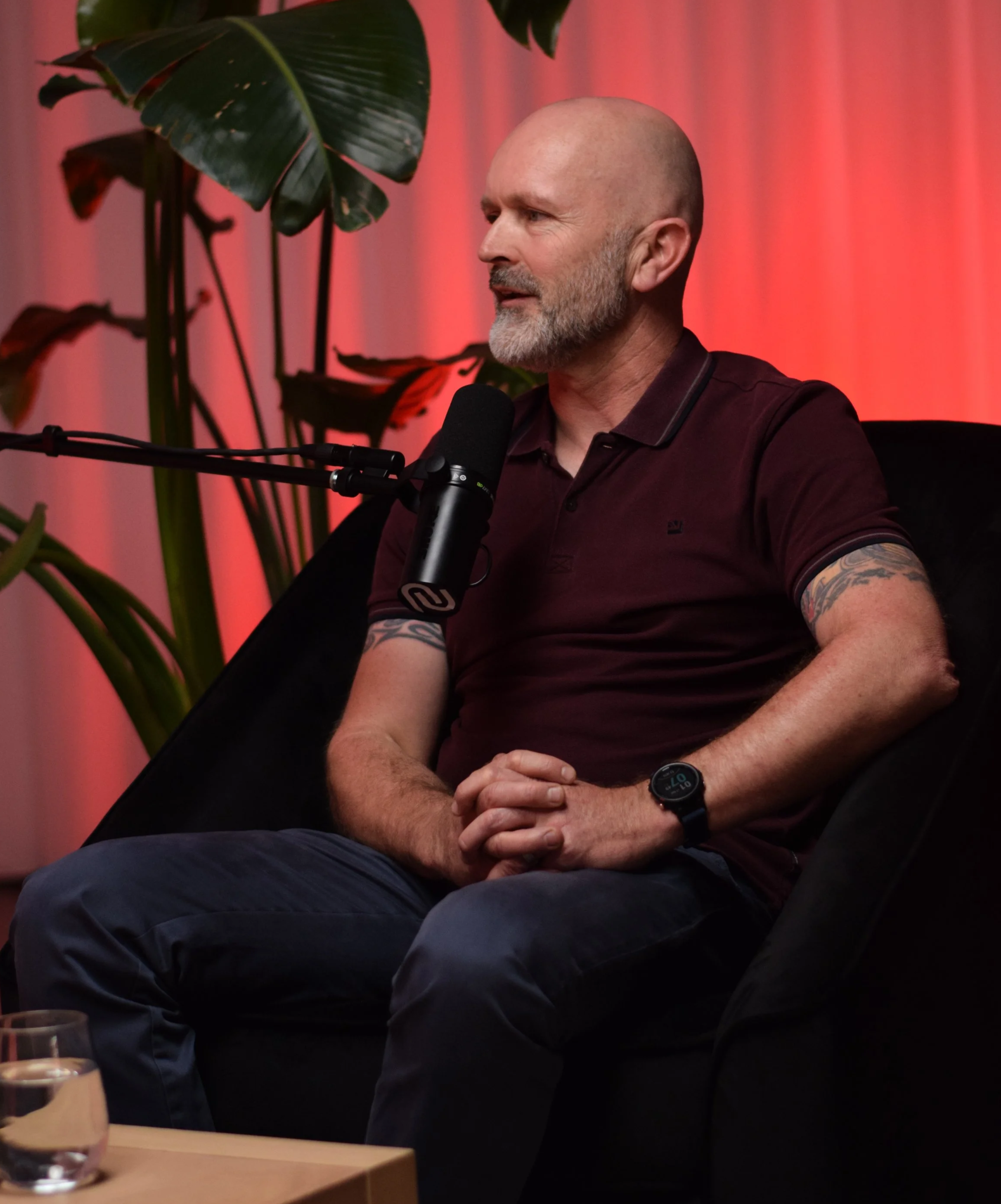 A man with a shaved head and beard, wearing a maroon polo shirt and a black smartwatch, sits on a black couch with his hands clasped, speaking into a microphone. There are large green plants and a pink curtain in the background.