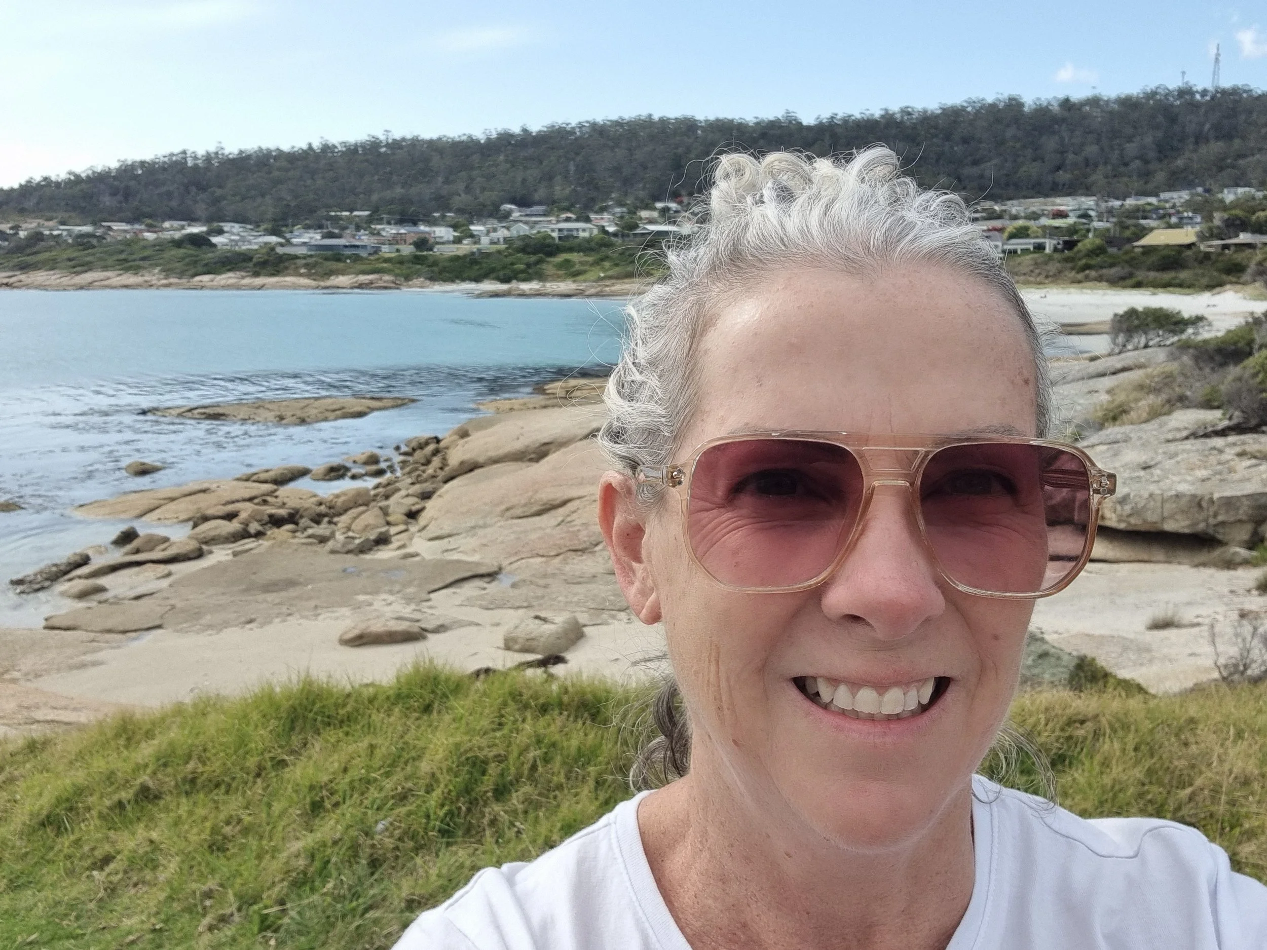 A woman with curly gray hair wearing pink-tinted glasses takes a selfie on a rocky beach with water, sand, and a hillside with houses in the background.