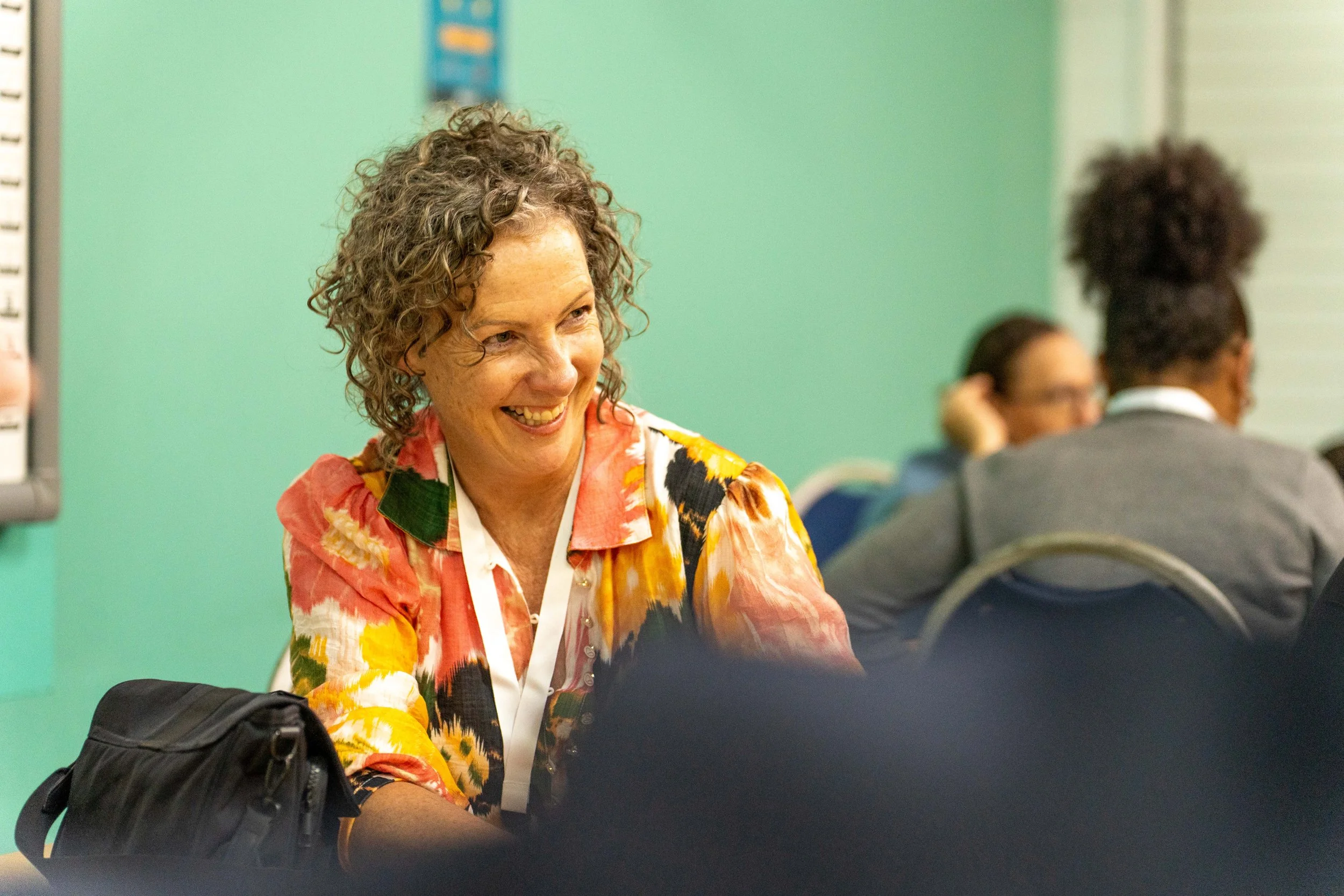 Woman with curly hair smiling and talking, seated at a table in a meeting or conference room.