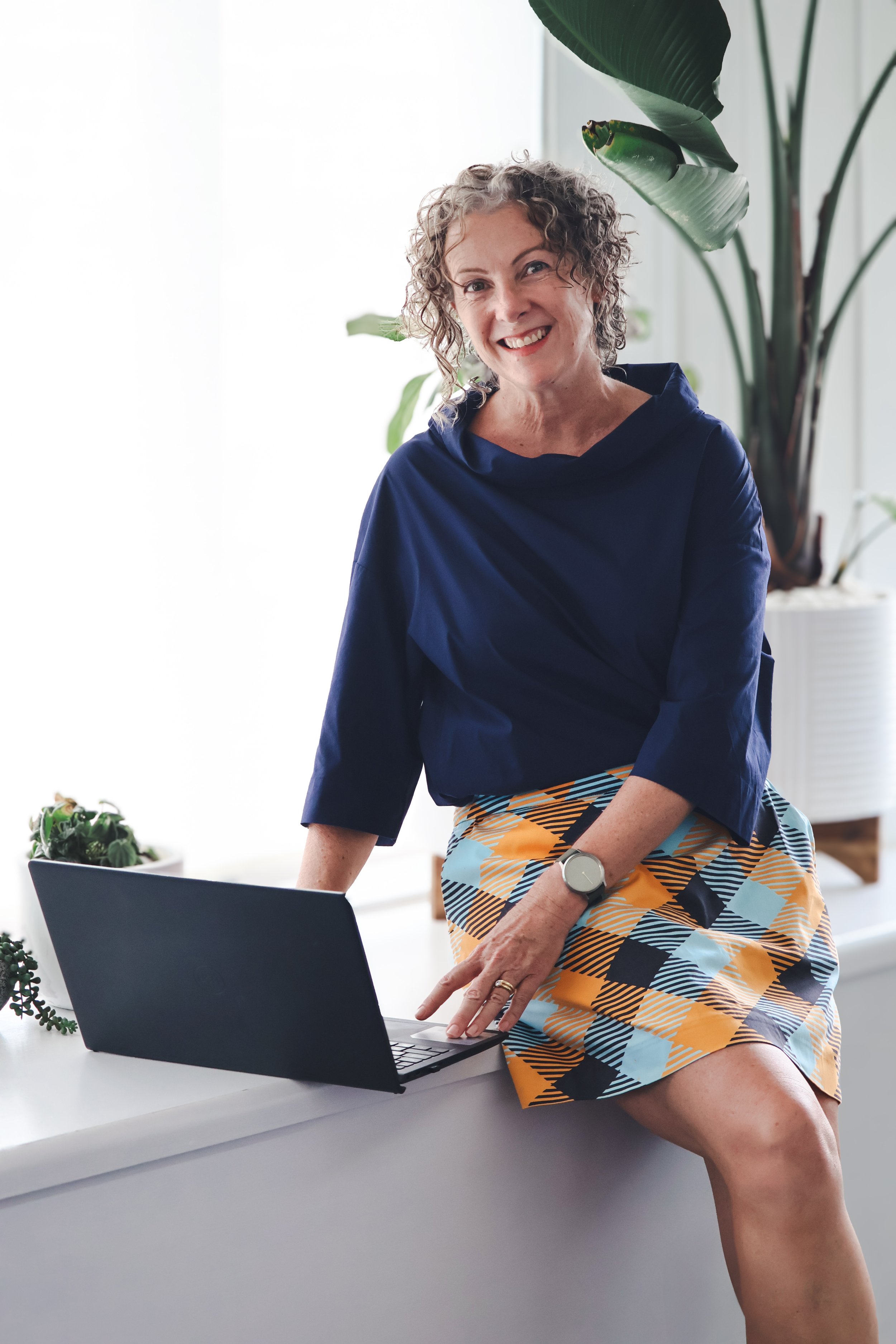 A woman with curly gray hair smiling while leaning on a laptop on a white desk, surrounded by green plants in a bright, airy room.