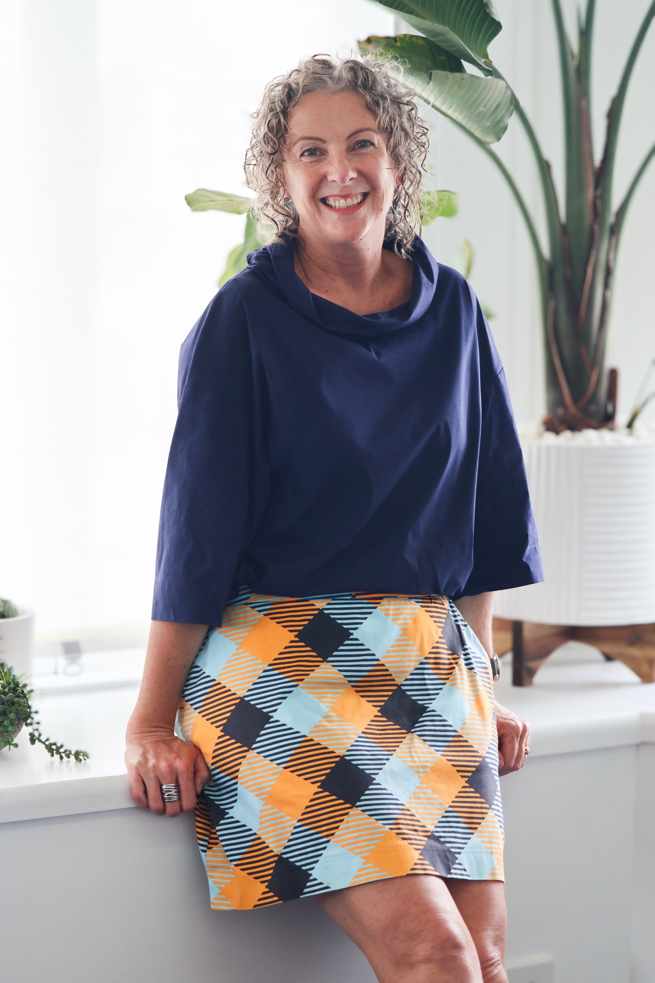 A woman with curly hair smiling, wearing a navy blue top and a colorful plaid skirt, sitting on a white surface with indoor plants in the background.