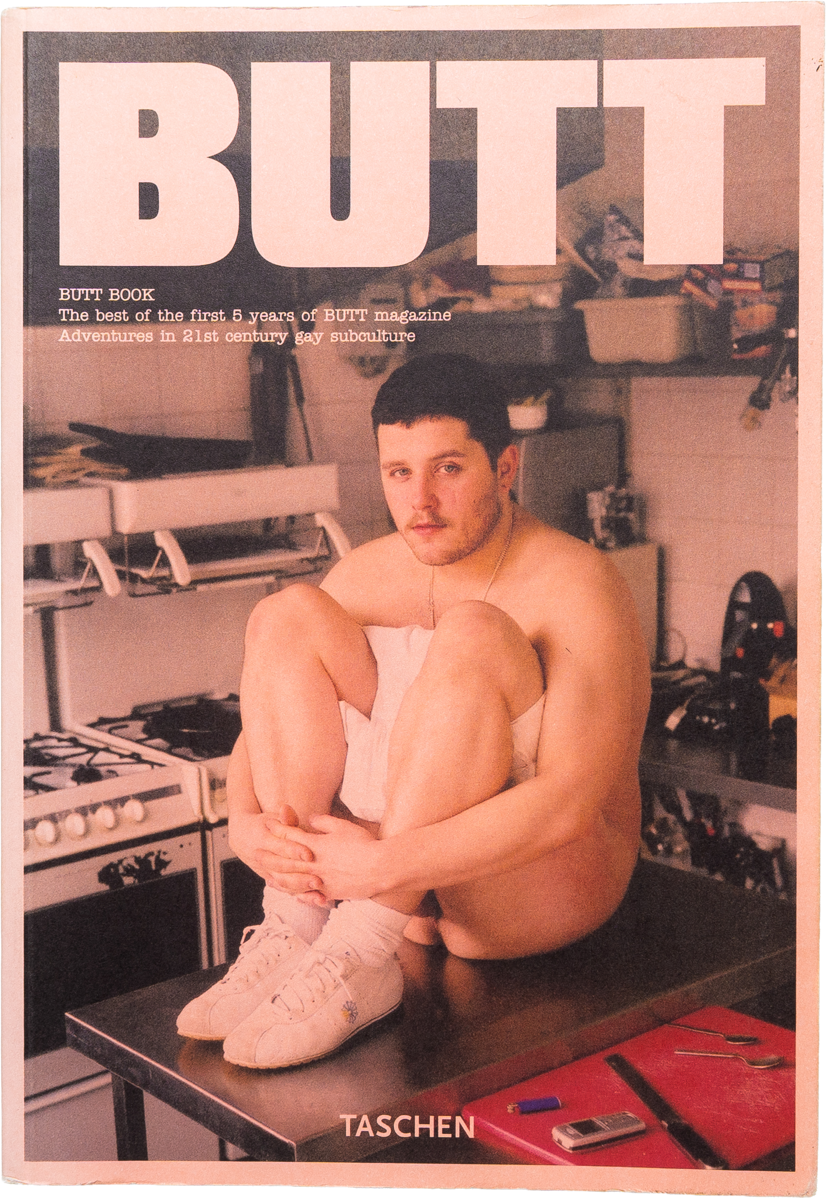 Cover of a magazine titled "BUTT" featuring a shirtless man sitting on a kitchen counter with his knees drawn to his chest, wearing white sneakers and socks, with kitchen appliances and shelves in the background.