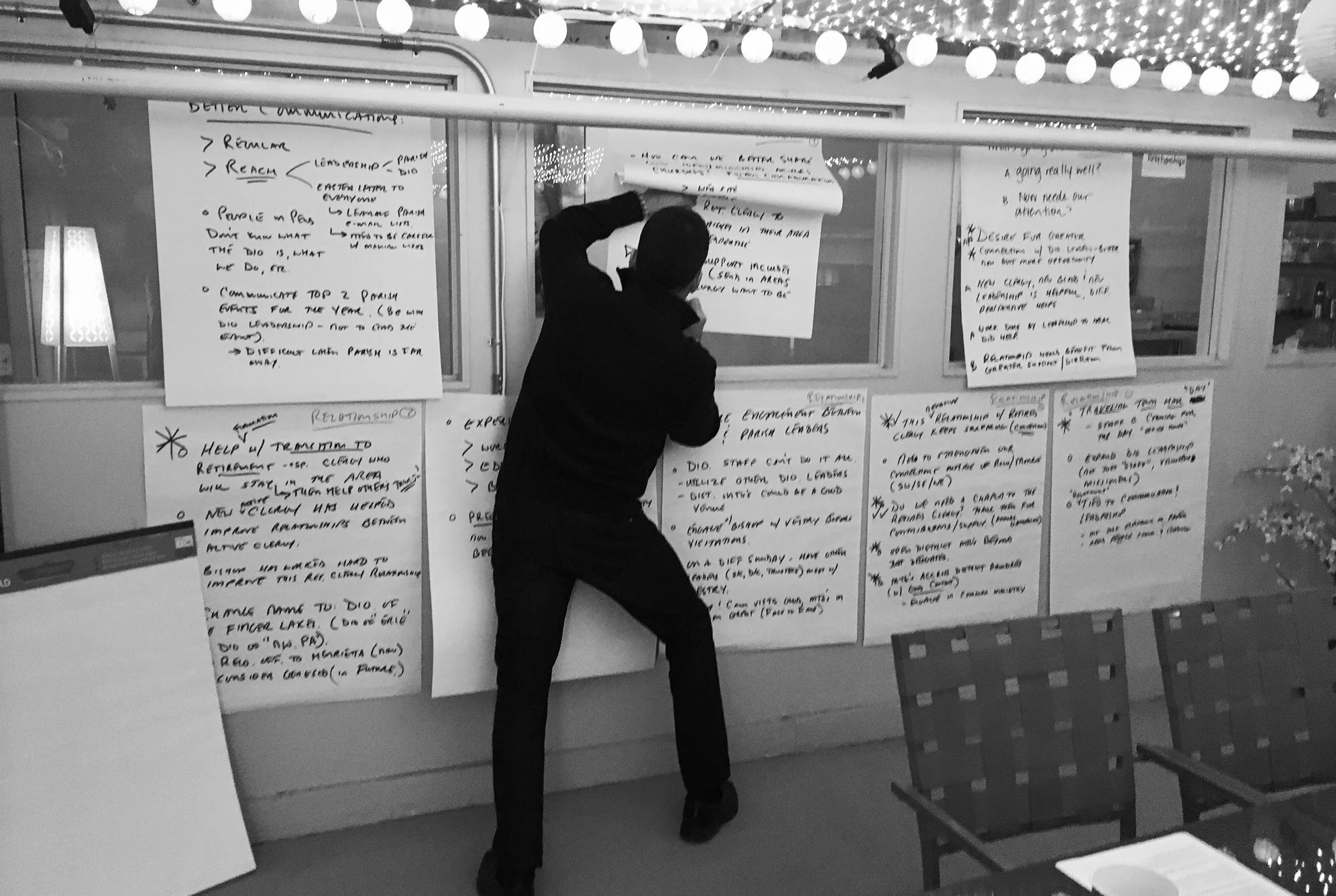 A person writing on large sheets of paper displayed on a glass window during a meeting or workshop, with string lights hanging above.