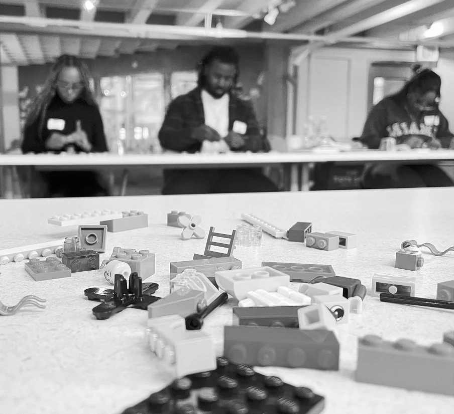 Scattered LEGO pieces on a table with three people in the background, sitting at a counter or table in a workshop or classroom setting.