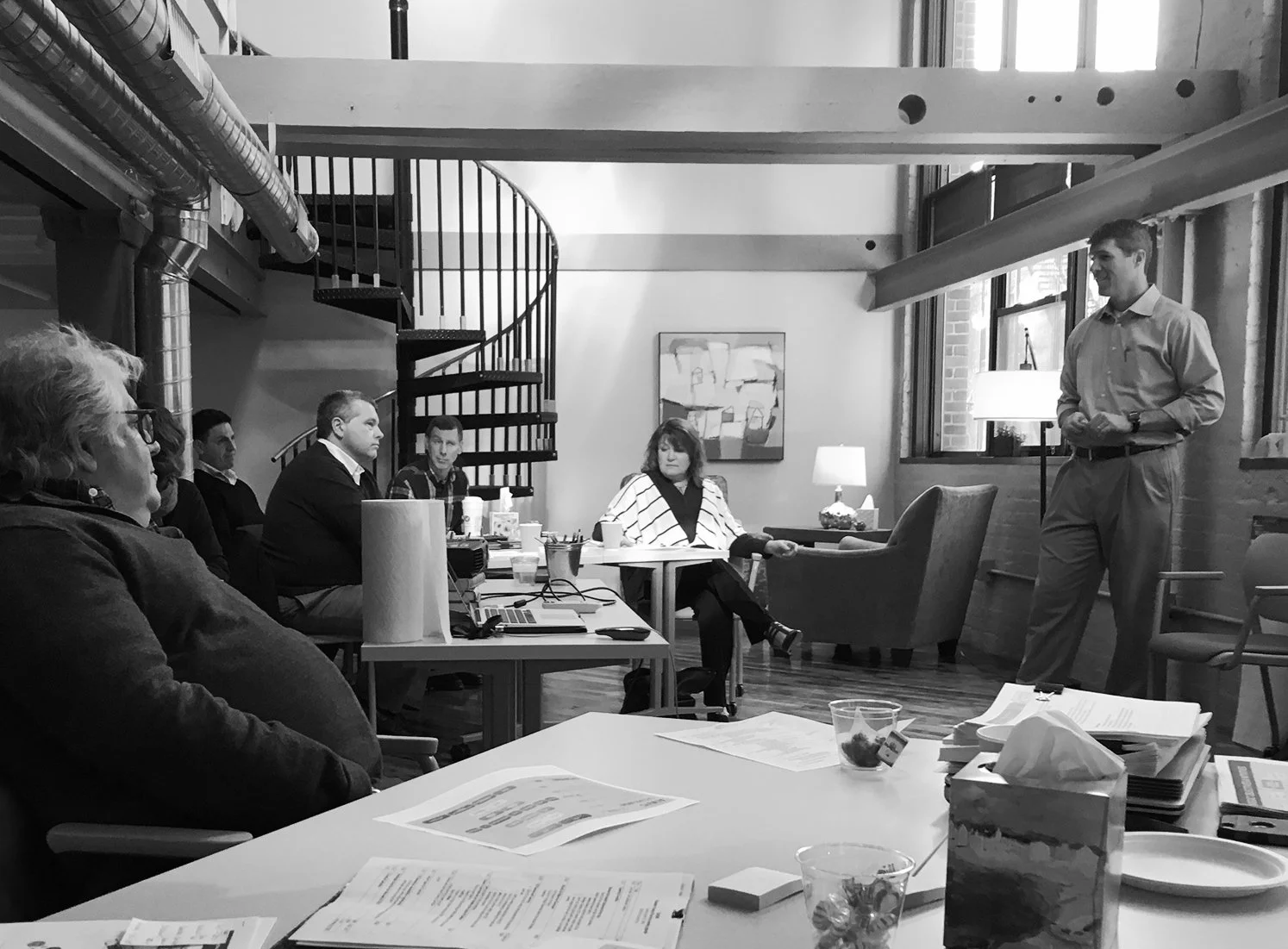 A group of people seated around a table in a conference room, listening to a standing speaker on the right side of the image. The room has large windows, exposed brick walls, and a staircase in the background.