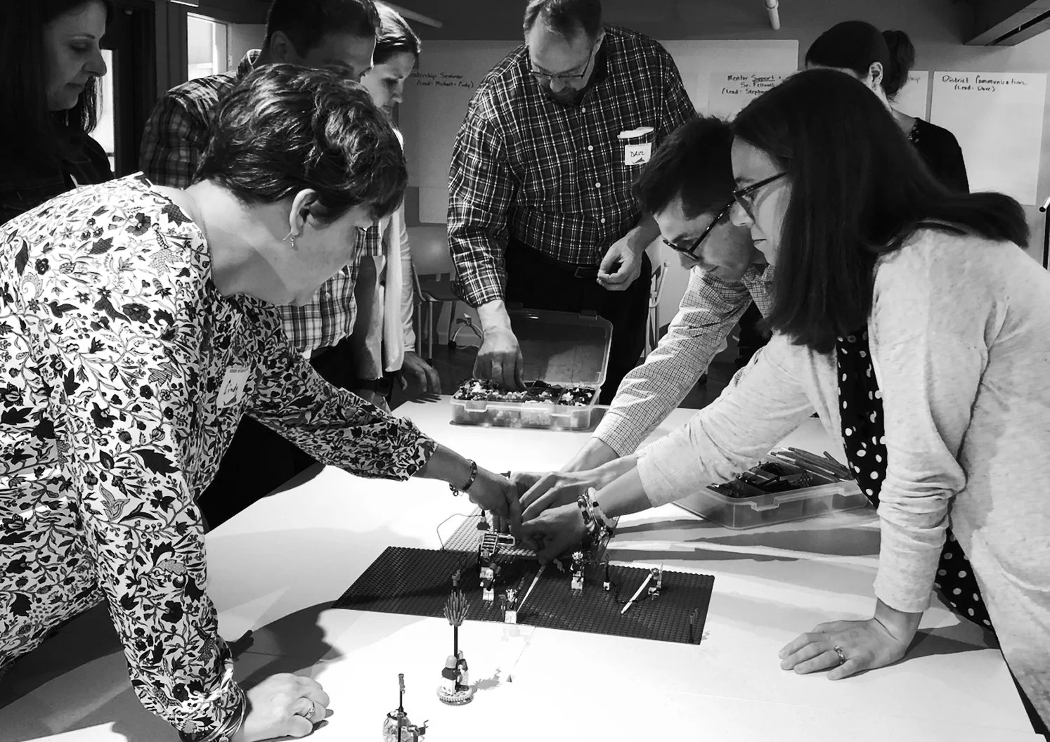 Group of adults gathered around a table, engaging in a collaborative activity with LEGO structures, in an indoor setting.