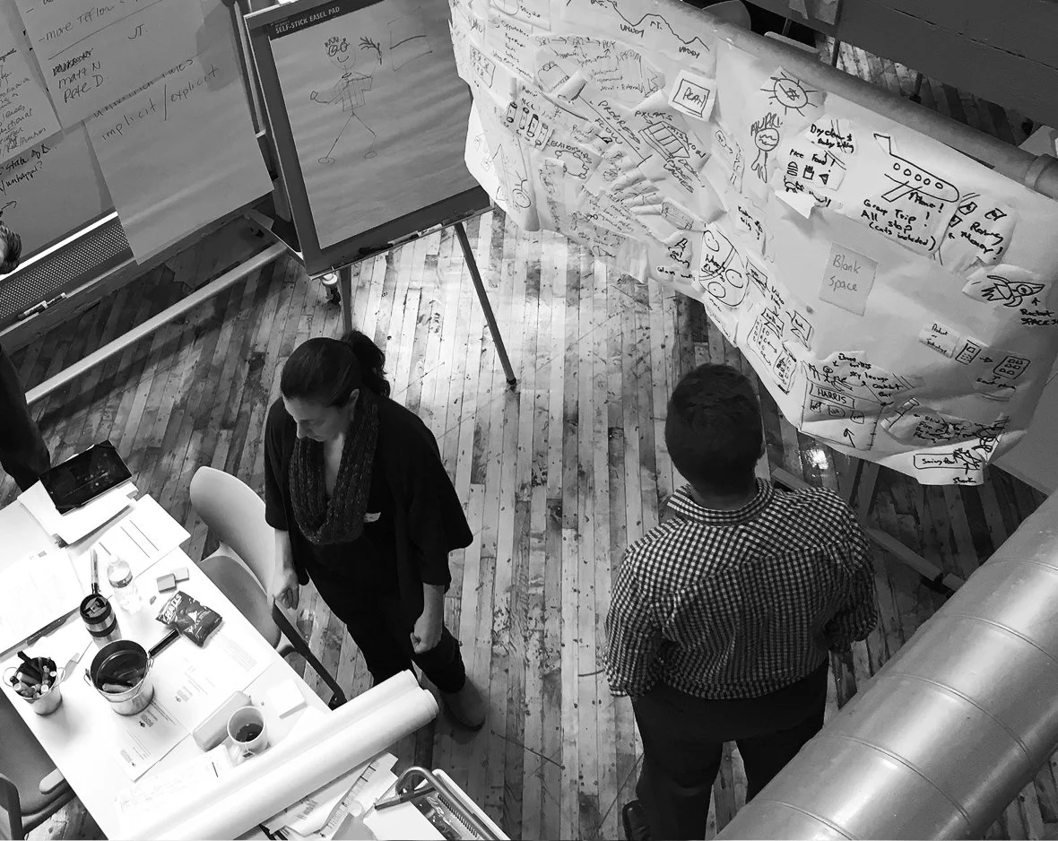 Two people in a workspace with large sheets of paper filled with sketches and notes, and a computer screen displaying a stick figure drawing.