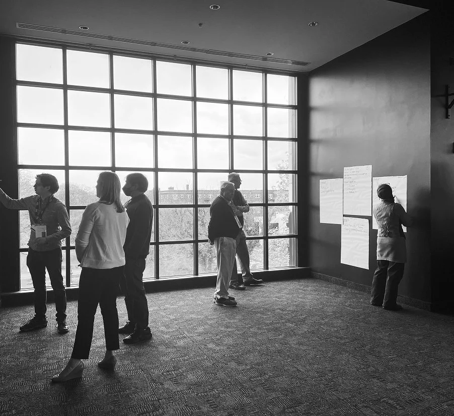 Six people in a modern conference room with large window, some discussing and writing on whiteboards.