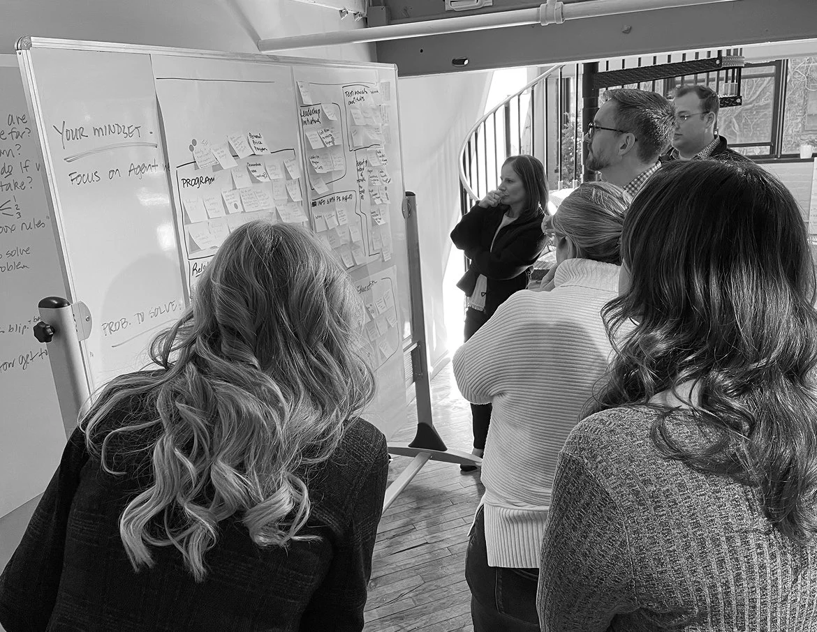 Group of people in a meeting room looking at a whiteboard covered with sticky notes and writing, discussing a project.