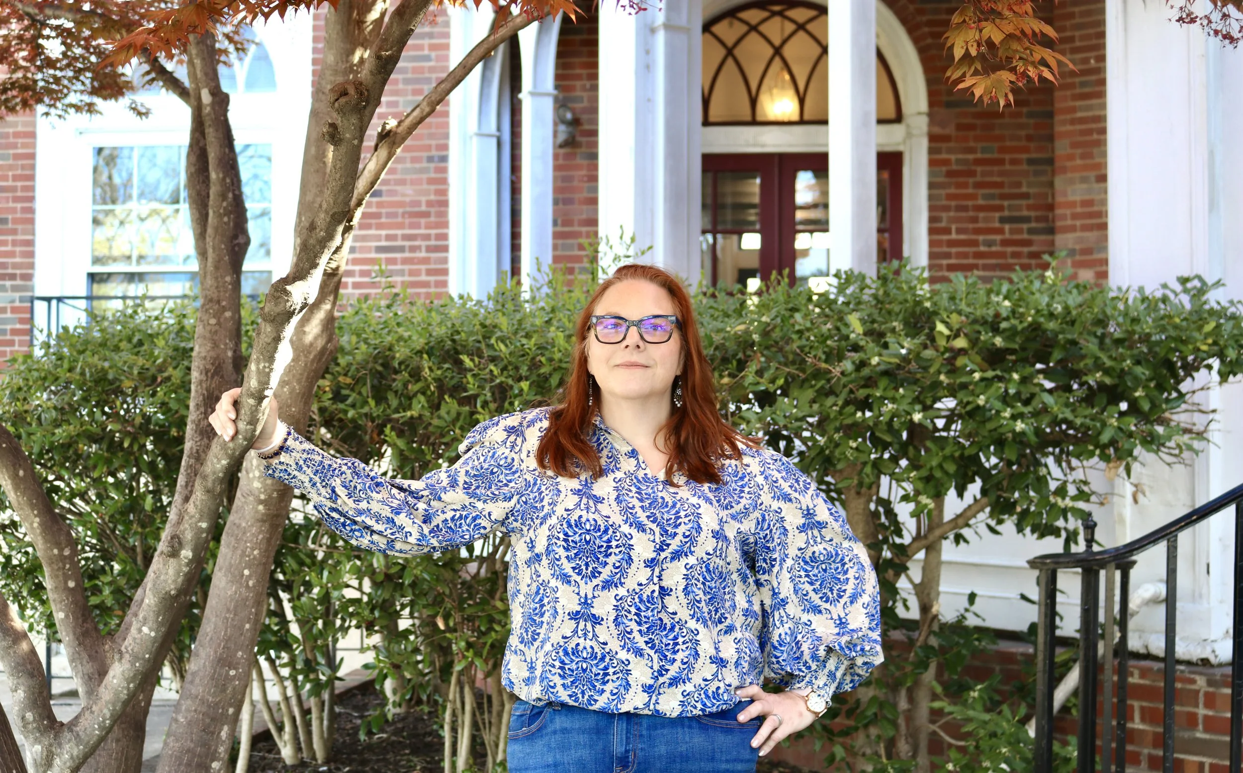 A woman with red hair and glasses standing outdoors in front of a shrub and a brick house, holding a tree branch and looking at the camera.