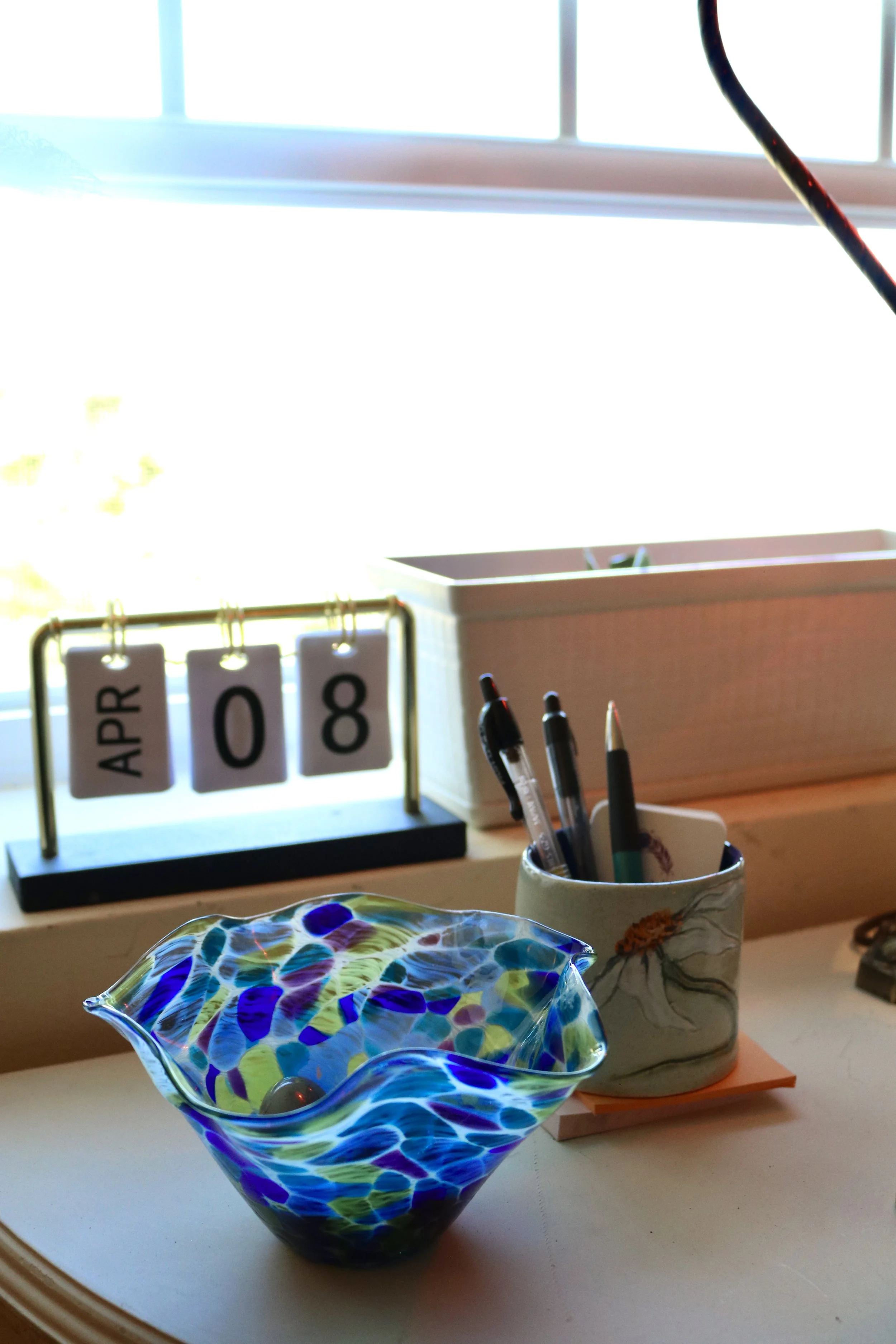 A desk with a colorful glass bowl, pens in a floral cup, a desk calendar showing April 8, and a wooden organizer near a window with sunlight.