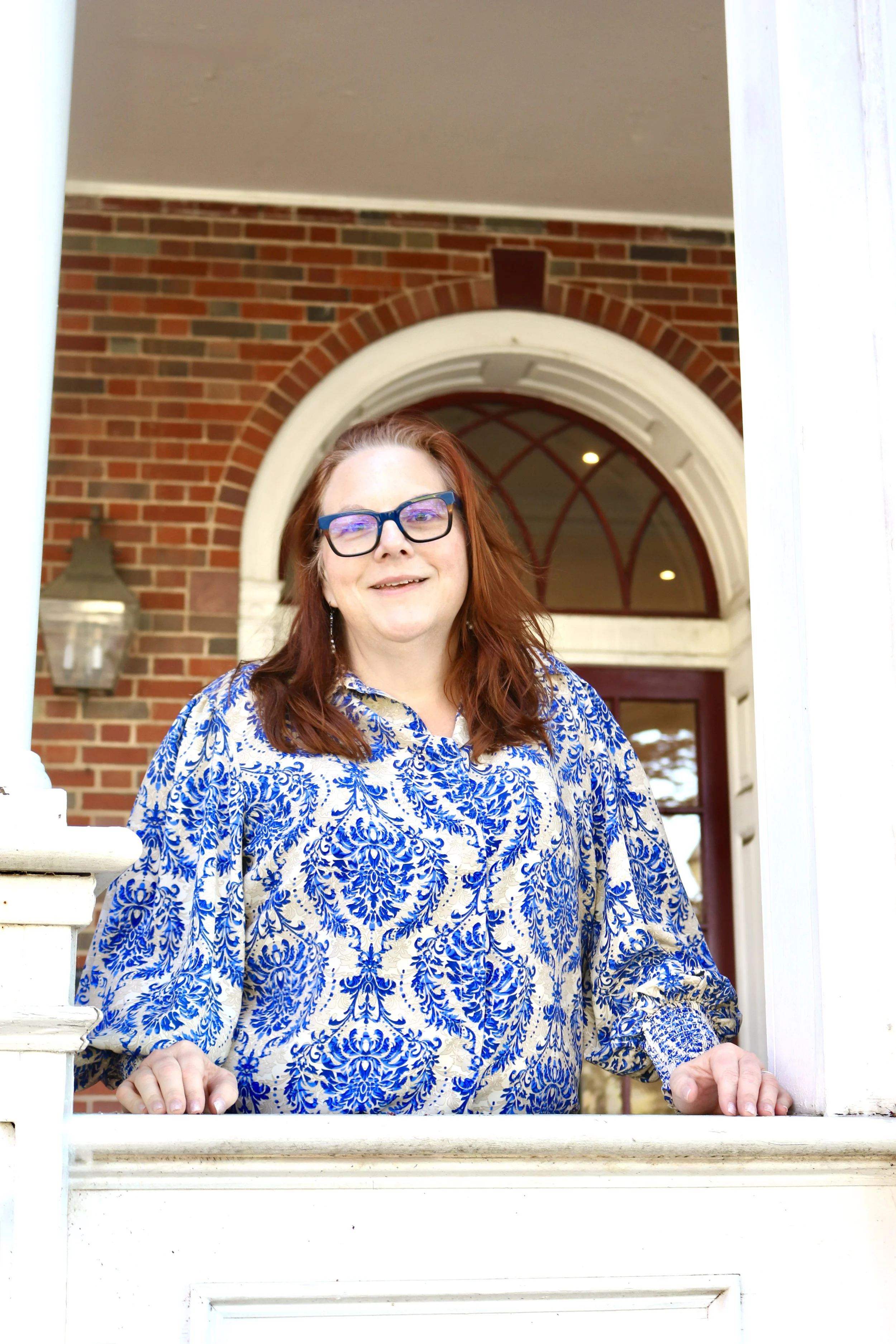 A woman with red hair and glasses standing on a porch in front of a brick house, smiling and looking at the camera.