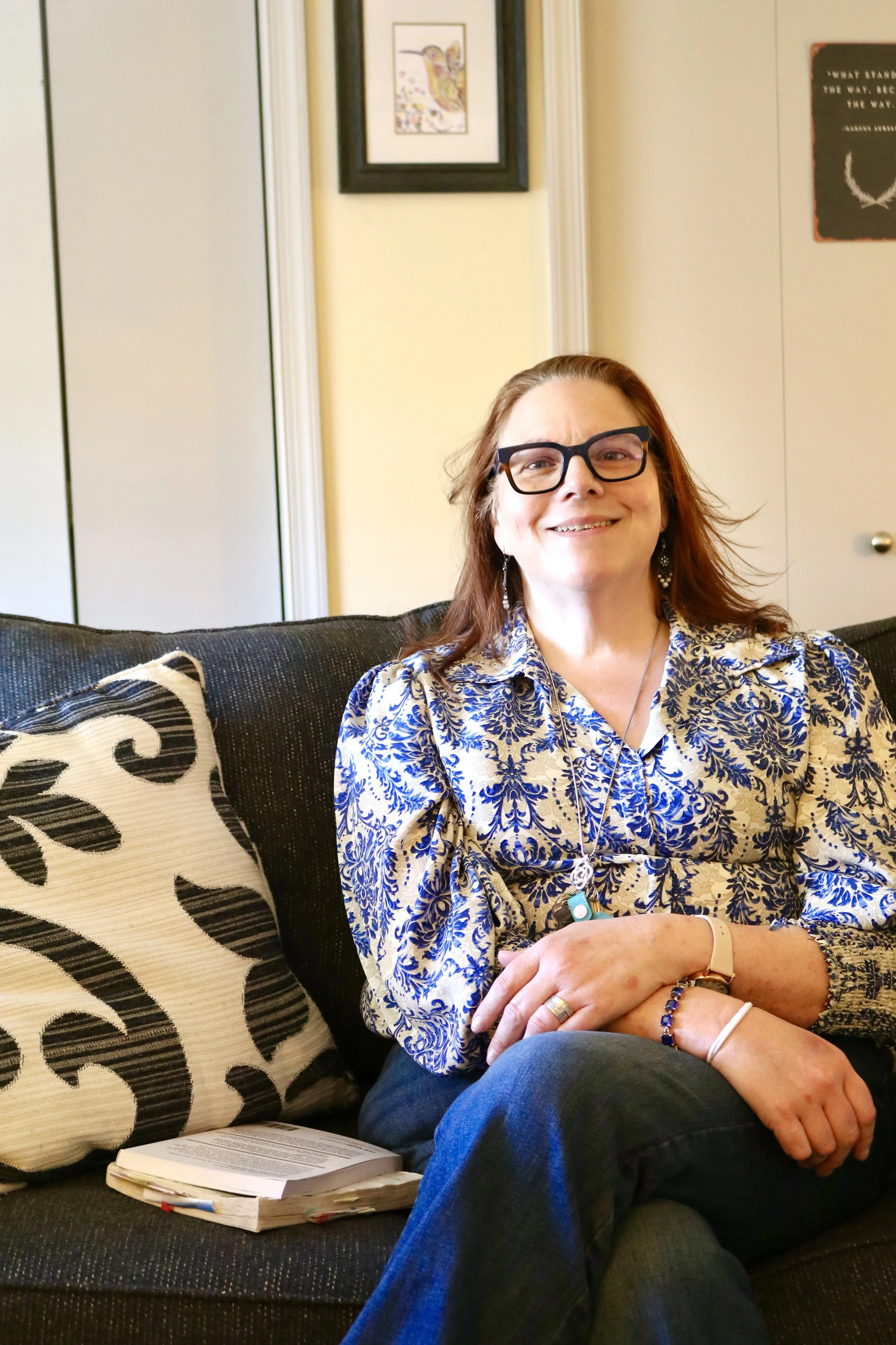 A woman with glasses, wearing a patterned blouse, sitting on a couch with black and white pillows, smiling at the camera. There are framed artworks and a door in the background.