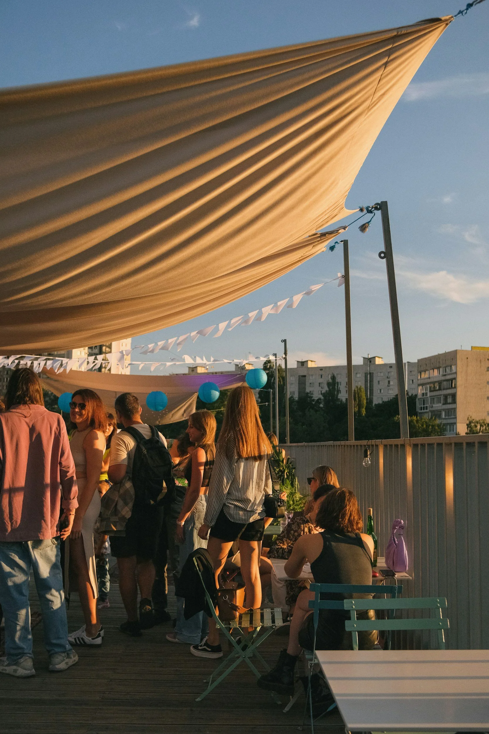 Reunión social al aire libre con personas conversando y disfrutando en una terraza decorada con manteles, globos azules y guirnaldas, durante una tarde con cielo despejado.