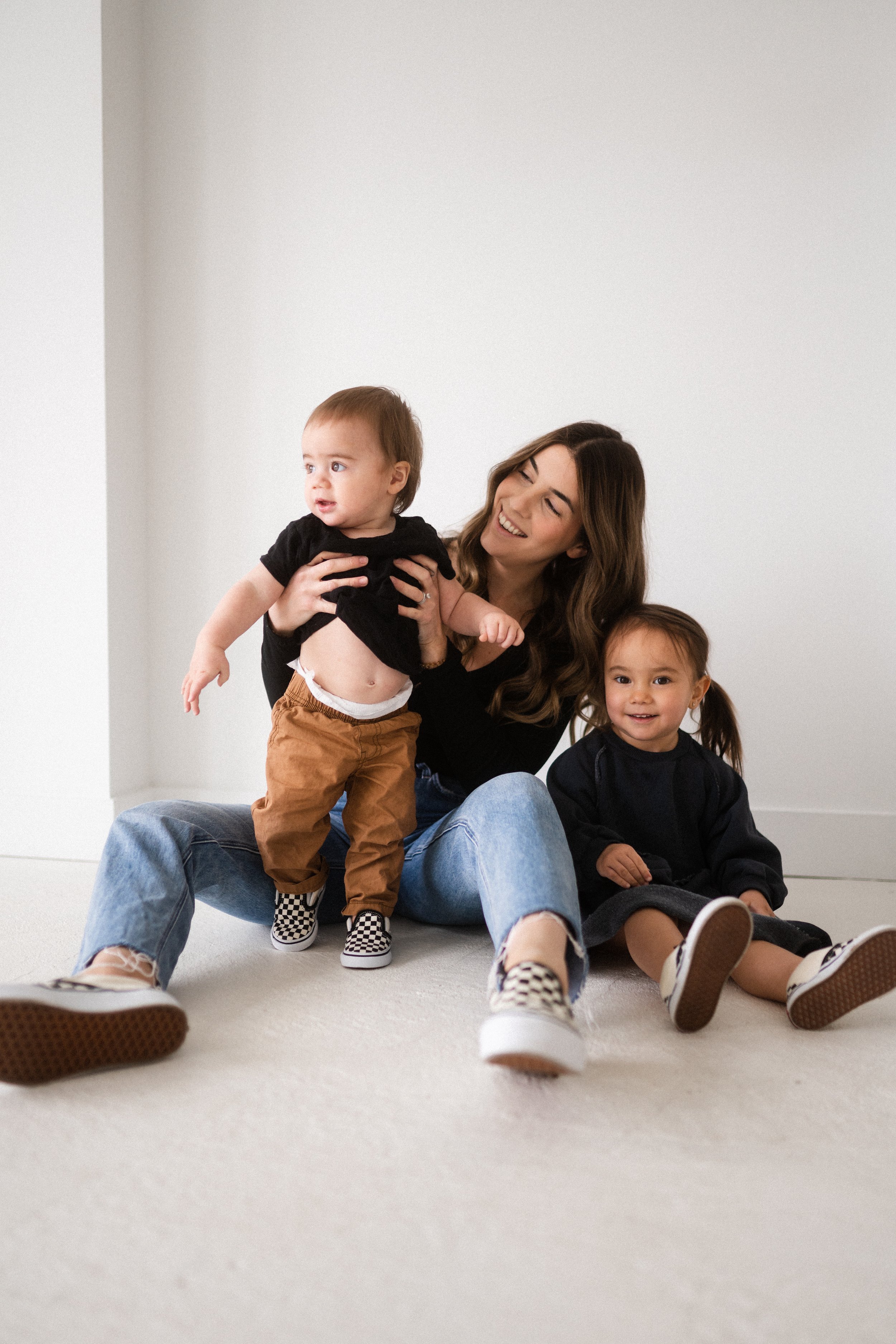 A woman with two young children sitting on a light-colored carpeted floor against a plain white wall. The woman is smiling, with the children sitting near her, one on her lap and one beside her.