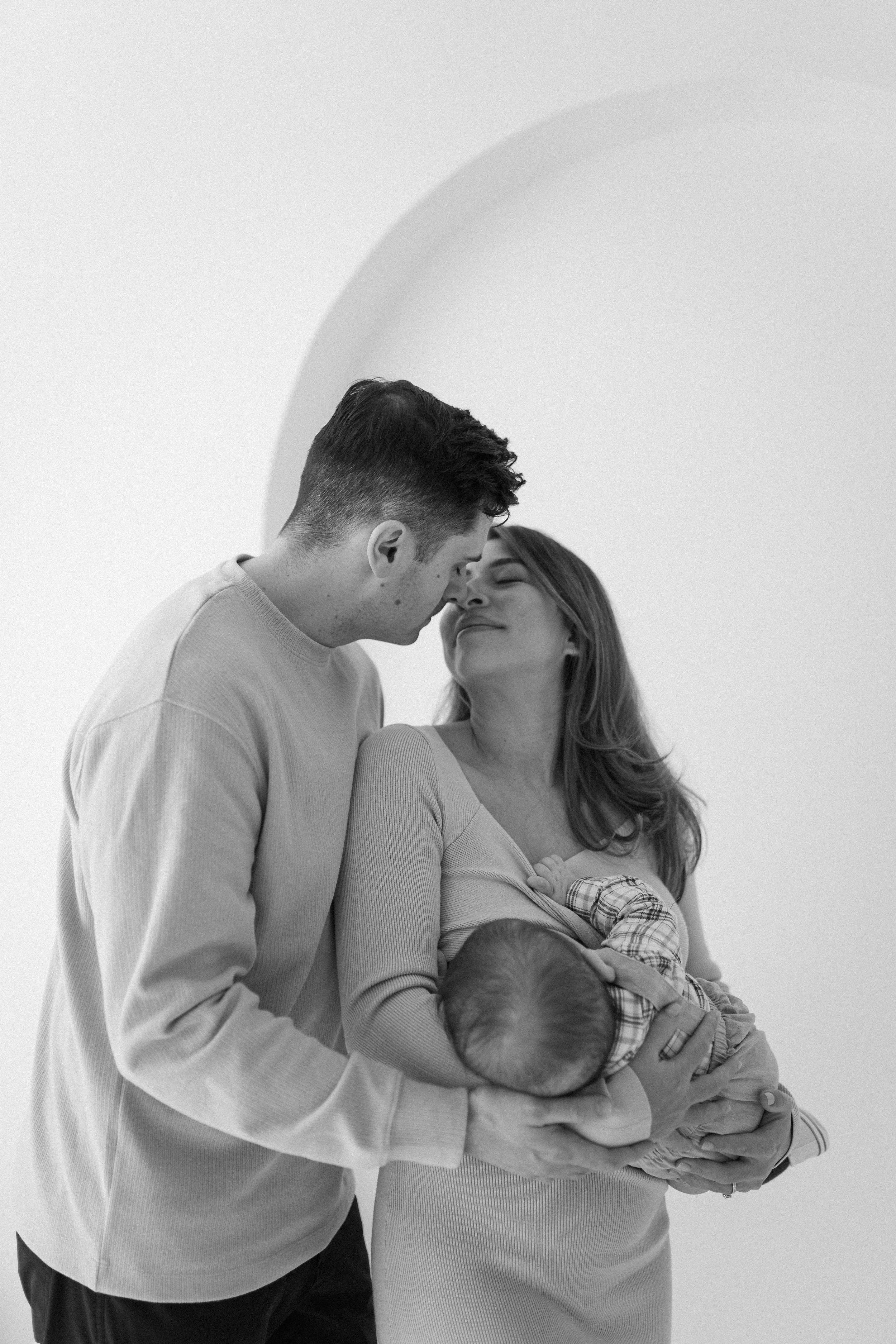 A black and white photo of a couple gently touching foreheads while holding a sleeping baby.