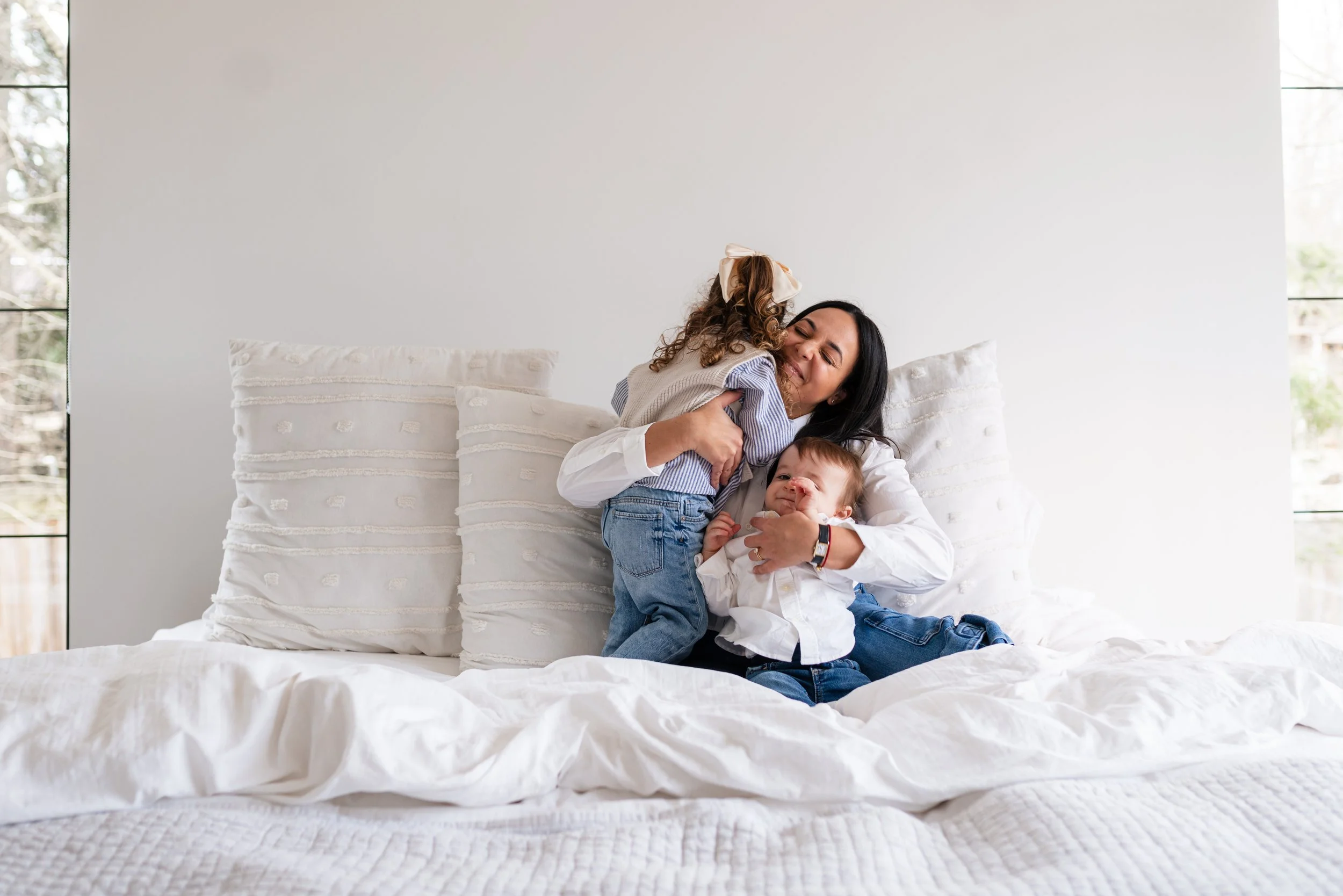 A woman sitting on a bed hugging two children, one boy and one girl, all smiling and enjoying a moment together in a bright bedroom with large windows.