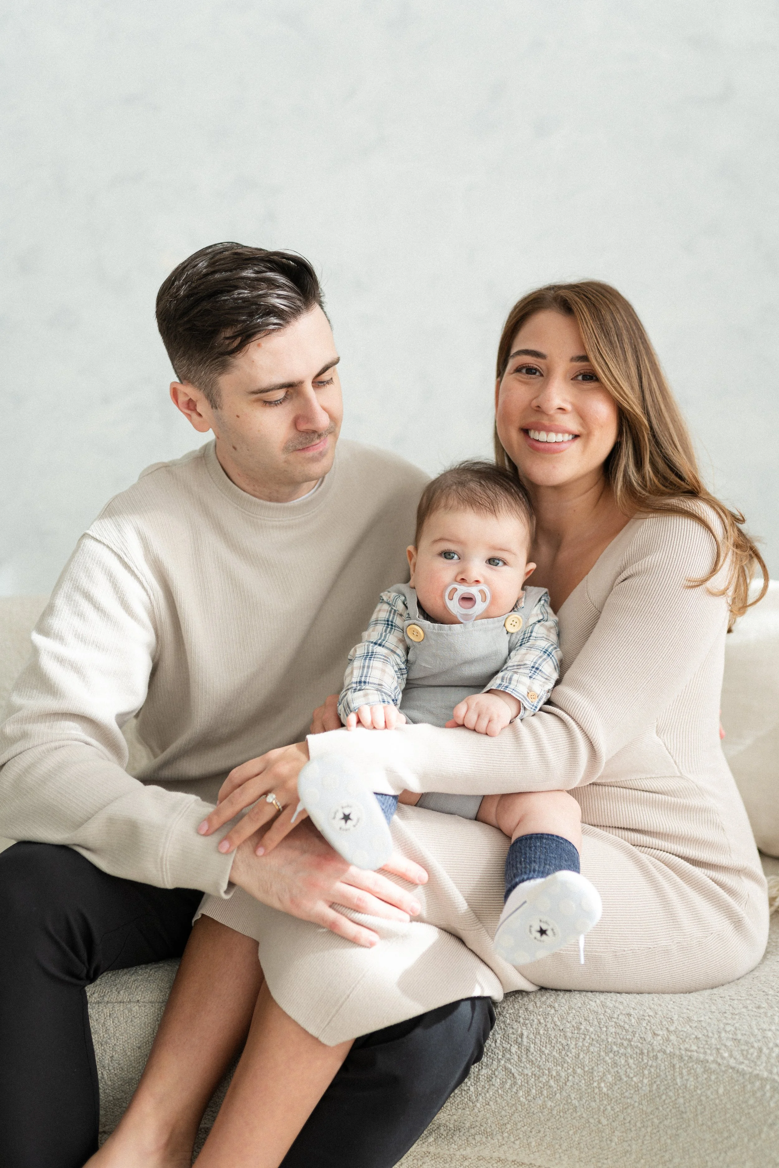 A smiling woman holding a baby with a pacifier, sitting on a couch with a man looking at the baby.