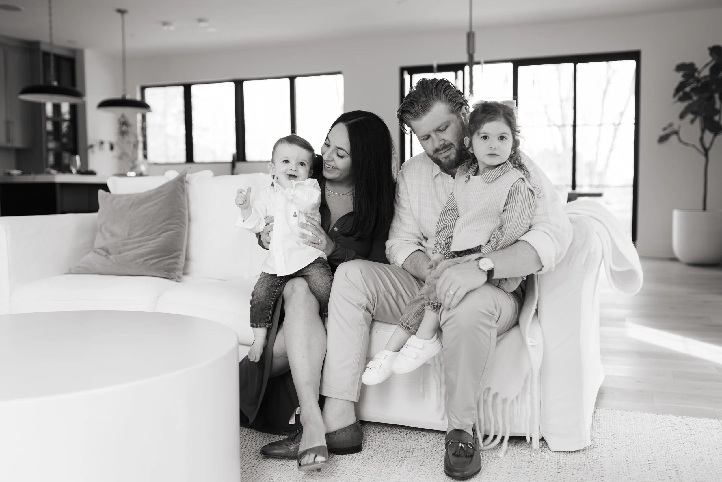 A family of four sitting on a sofa in a modern living room. The mother is holding a toddler, and the father is sitting with a young girl on his lap. All are smiling or looking content.