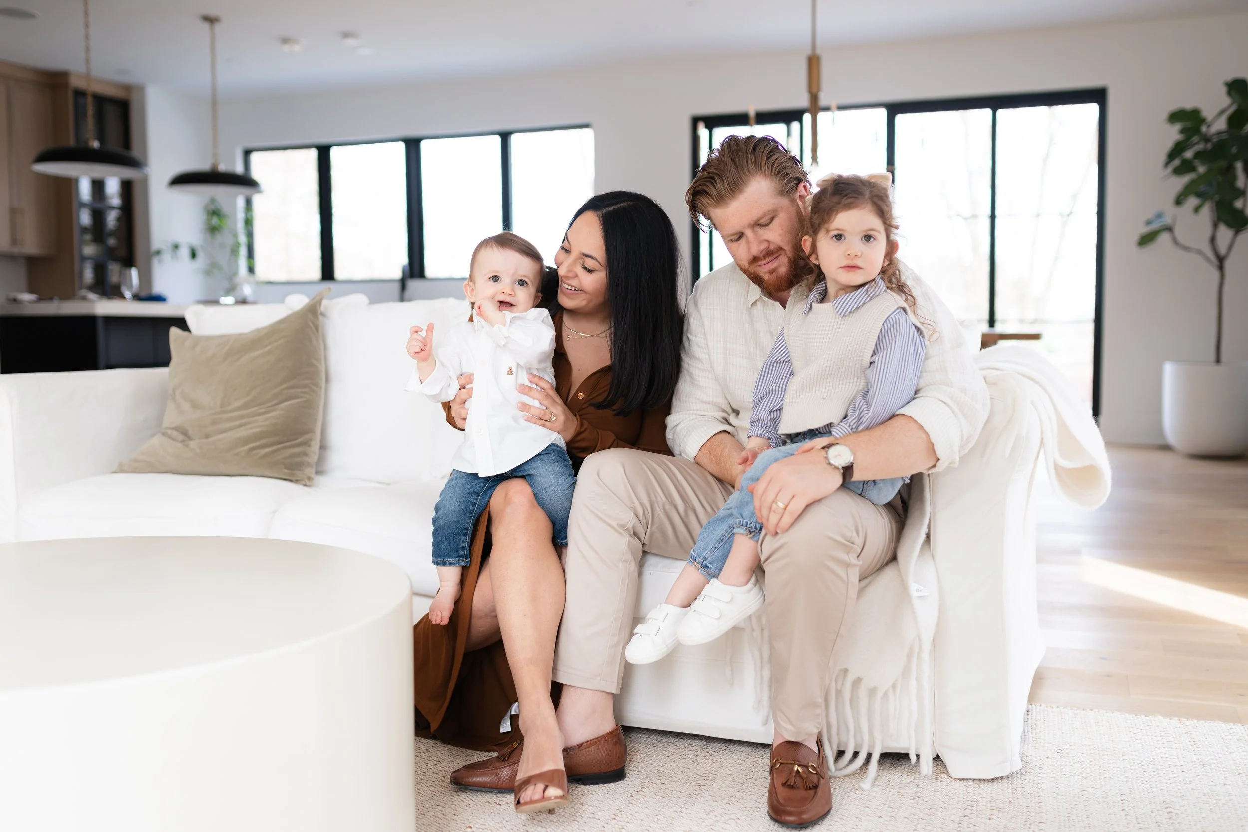 A family of four sitting on a white couch in a modern living room. The mother is holding a baby, and the father is holding a young girl. The room has large windows, a beige rug, and houseplants.