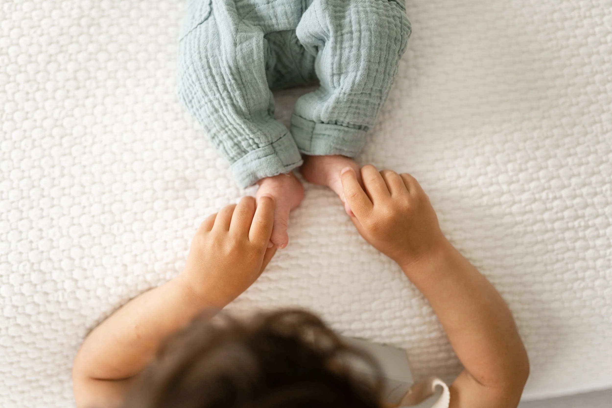 A child holding and gently pulling the feet of a newborn baby dressed in gray pajamas on a textured cream-colored blanket.