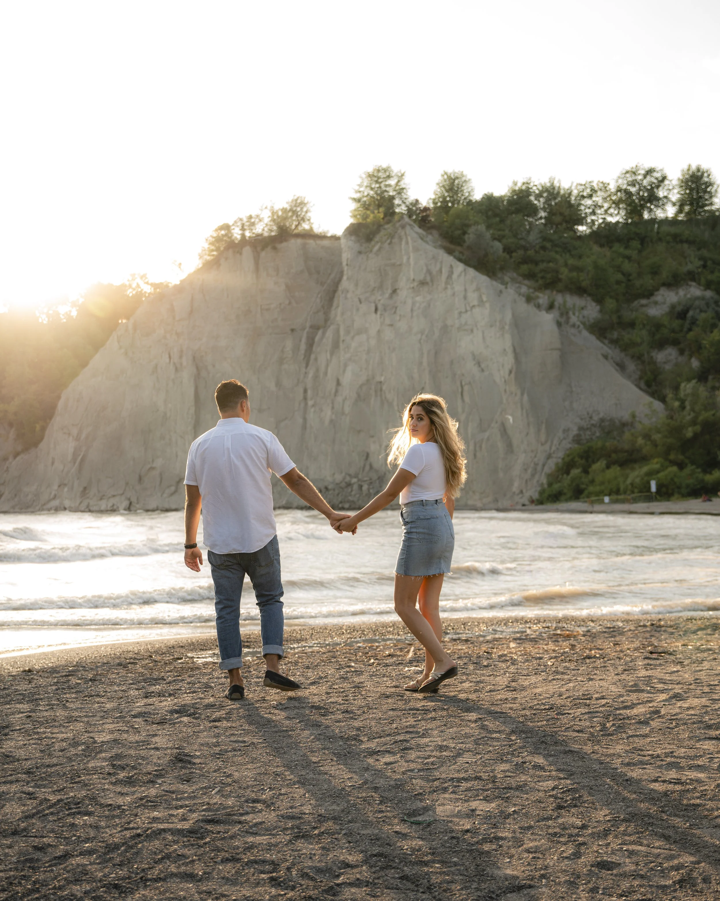A young couple holding hands on the beach during sunset with white cliffs and greenery in the background.