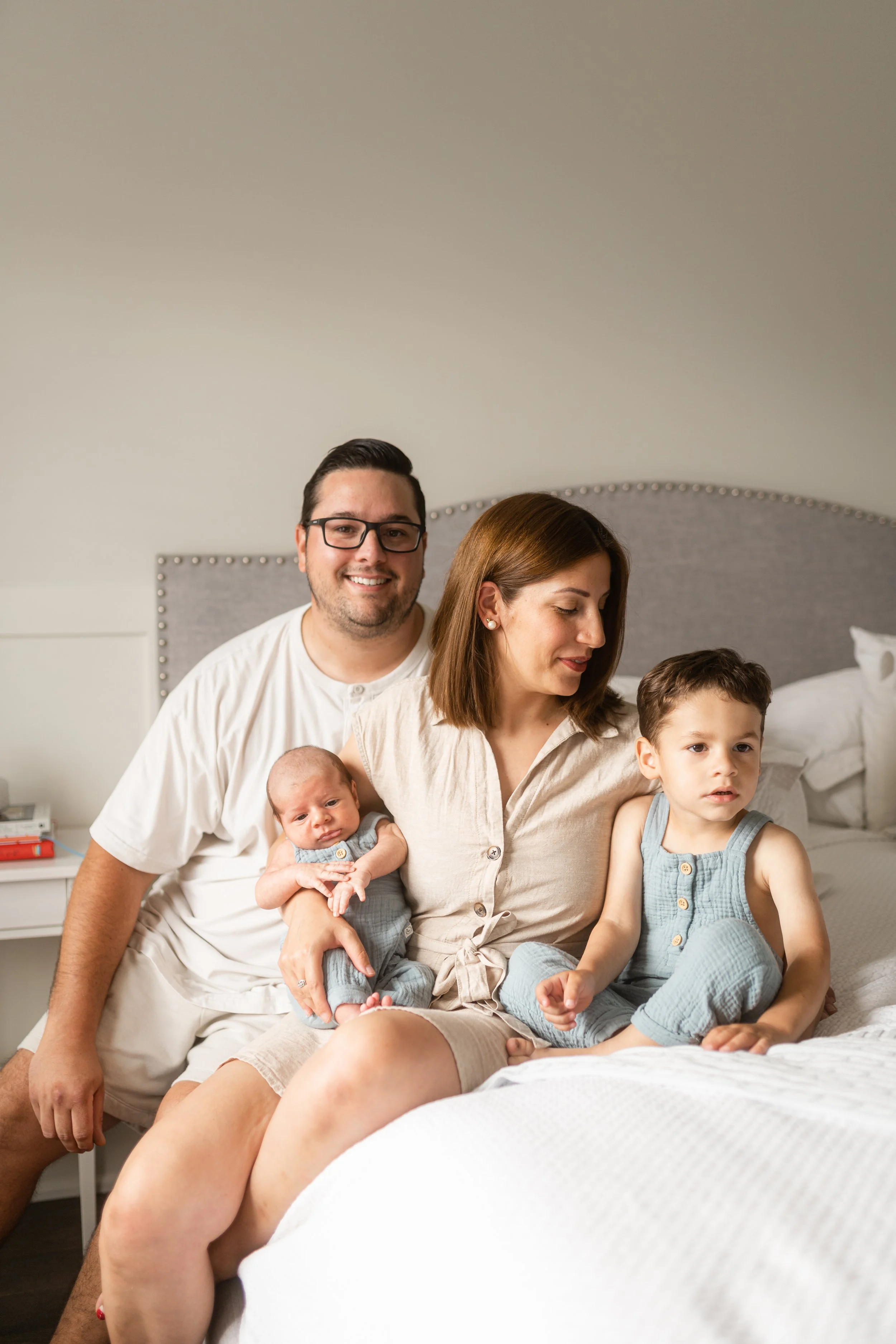 A family of four sitting on a bed in a bedroom. The father, mother, a young boy, and a newborn baby are present.