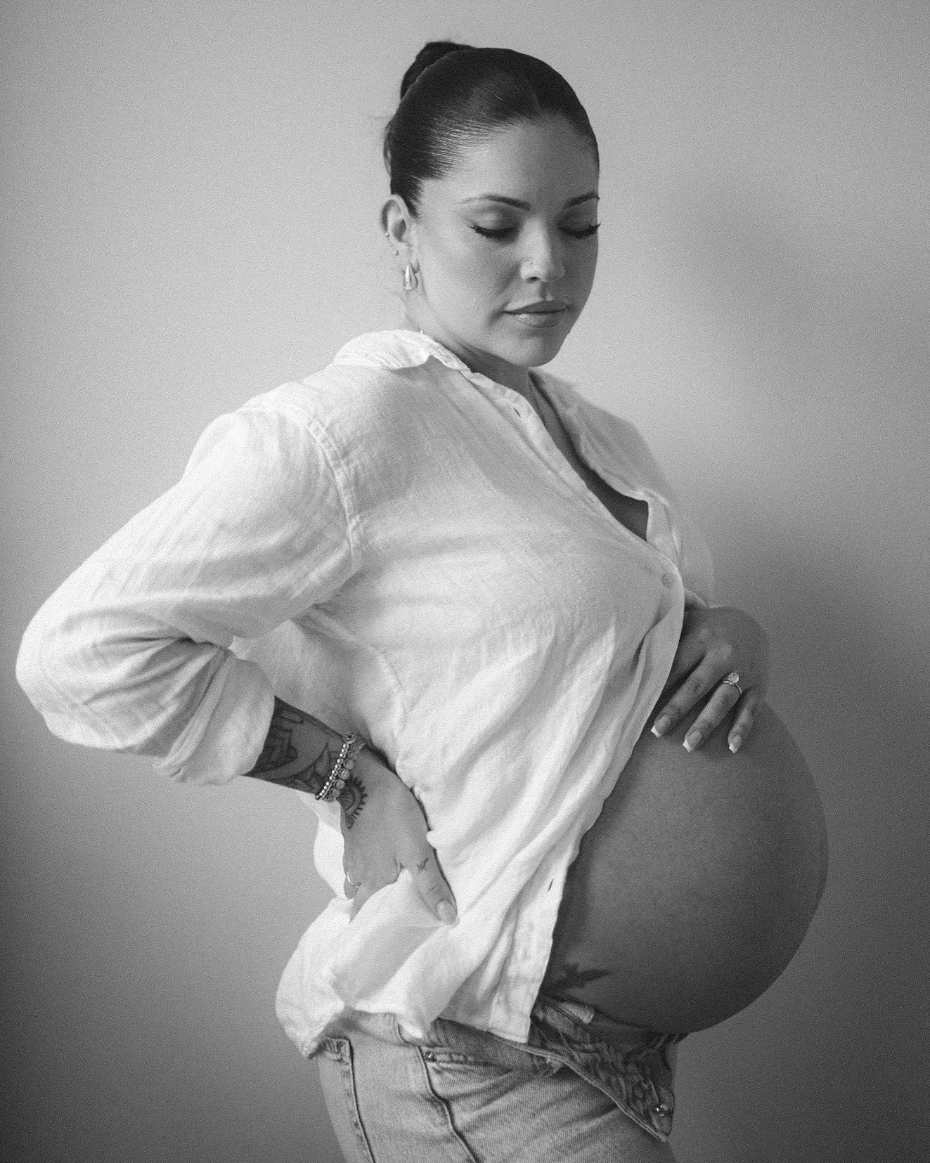 A pregnant woman with dark hair tied back, wearing a light-colored shirt with rolled-up sleeves, stands with her hand on her belly, looking down and to the side, against a plain background.
