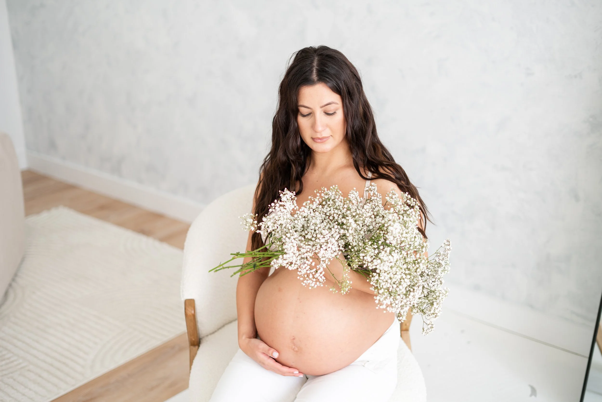A pregnant woman with long dark hair sitting on a white armchair, holding a large bouquet of white baby's breath flowers, and looking down at her belly.