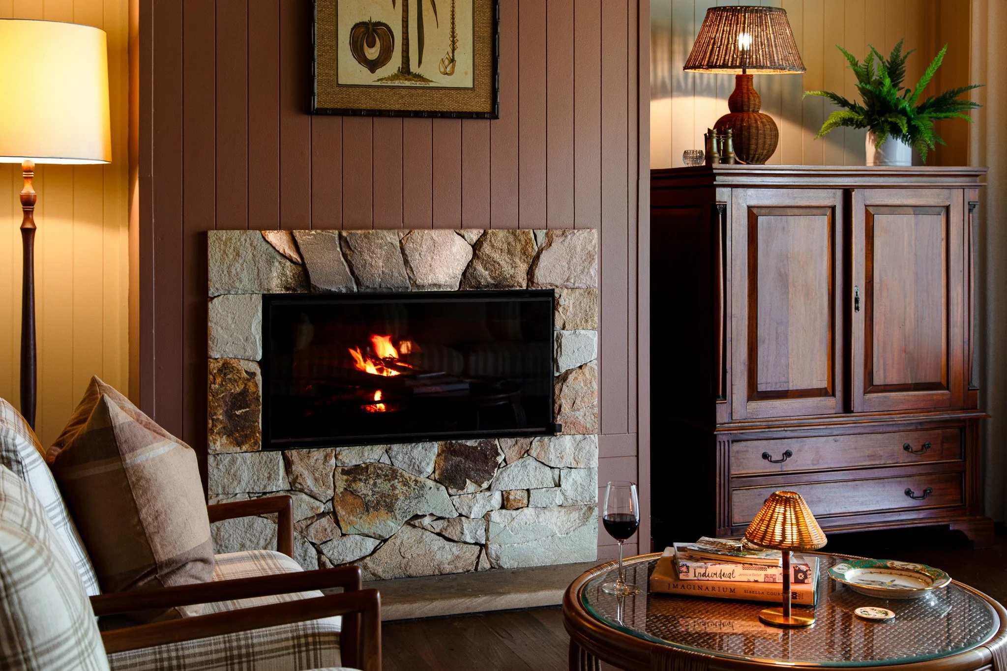 Cozy living room corner with a lit fireplace surrounded by rocks, a plaid armchair with cushions, a round glass-top coffee table with books, a small lamp, a glass of red wine, a wooden cabinet with a plant and lamp on top, a framed picture, and a tal