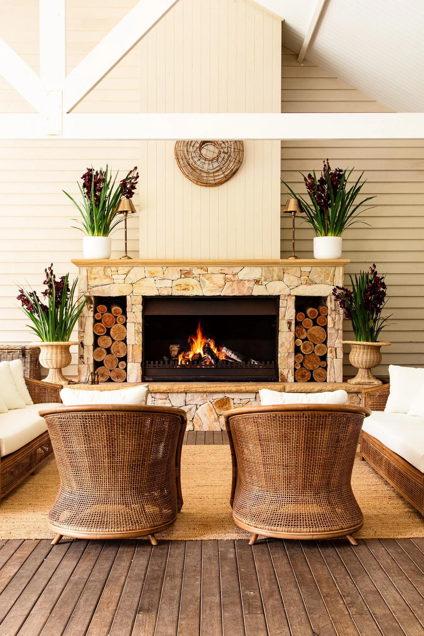 Cozy living room with a stone fireplace, wooden logs, potted plants, wicker chairs, white cushions, and a fire burning.