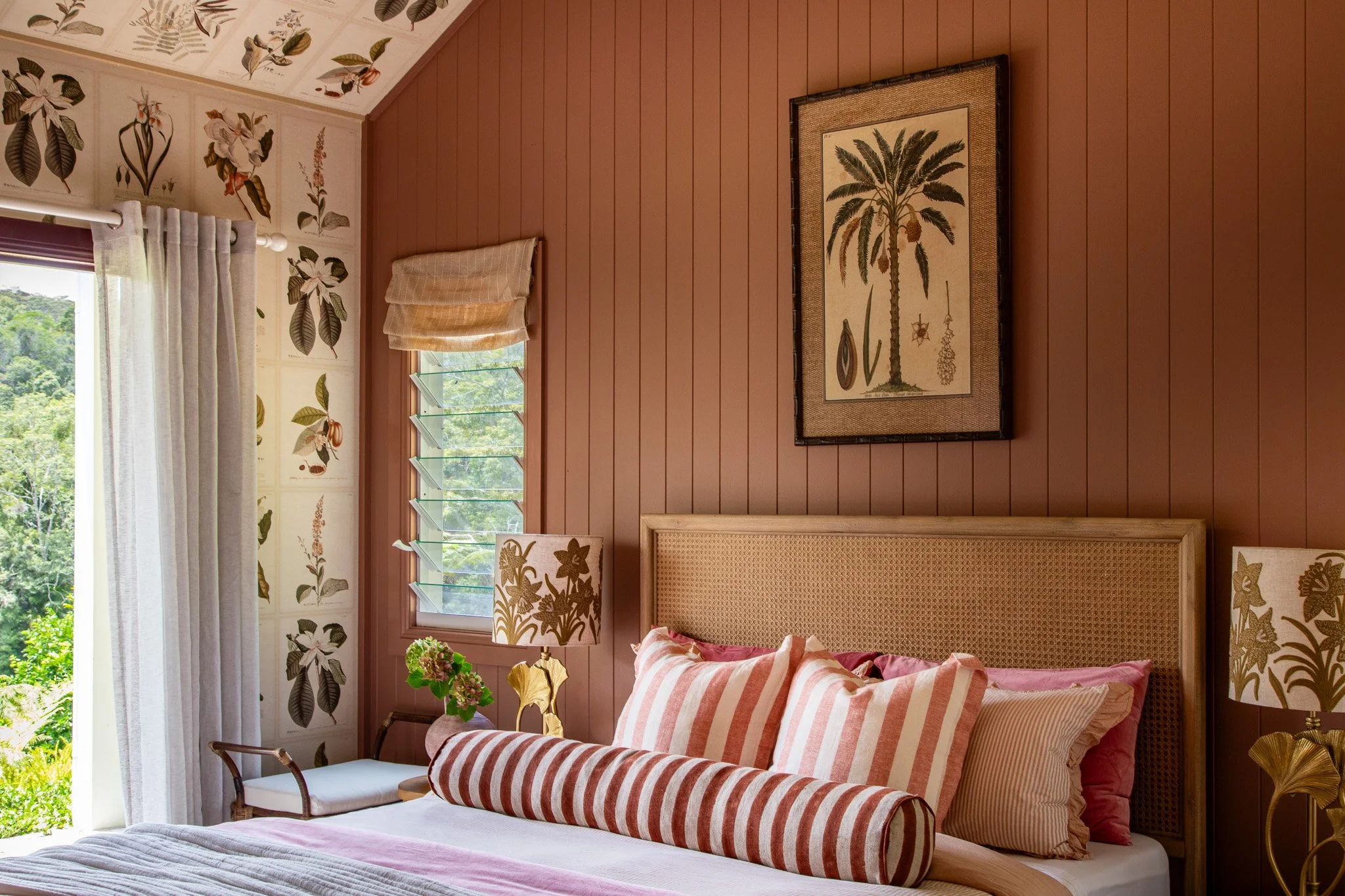 A cozy bedroom with pink and beige decor, featuring a bed with striped pillows, floral lamps, and botanical artwork on the walls. Window with white curtains and view of greenery outside.