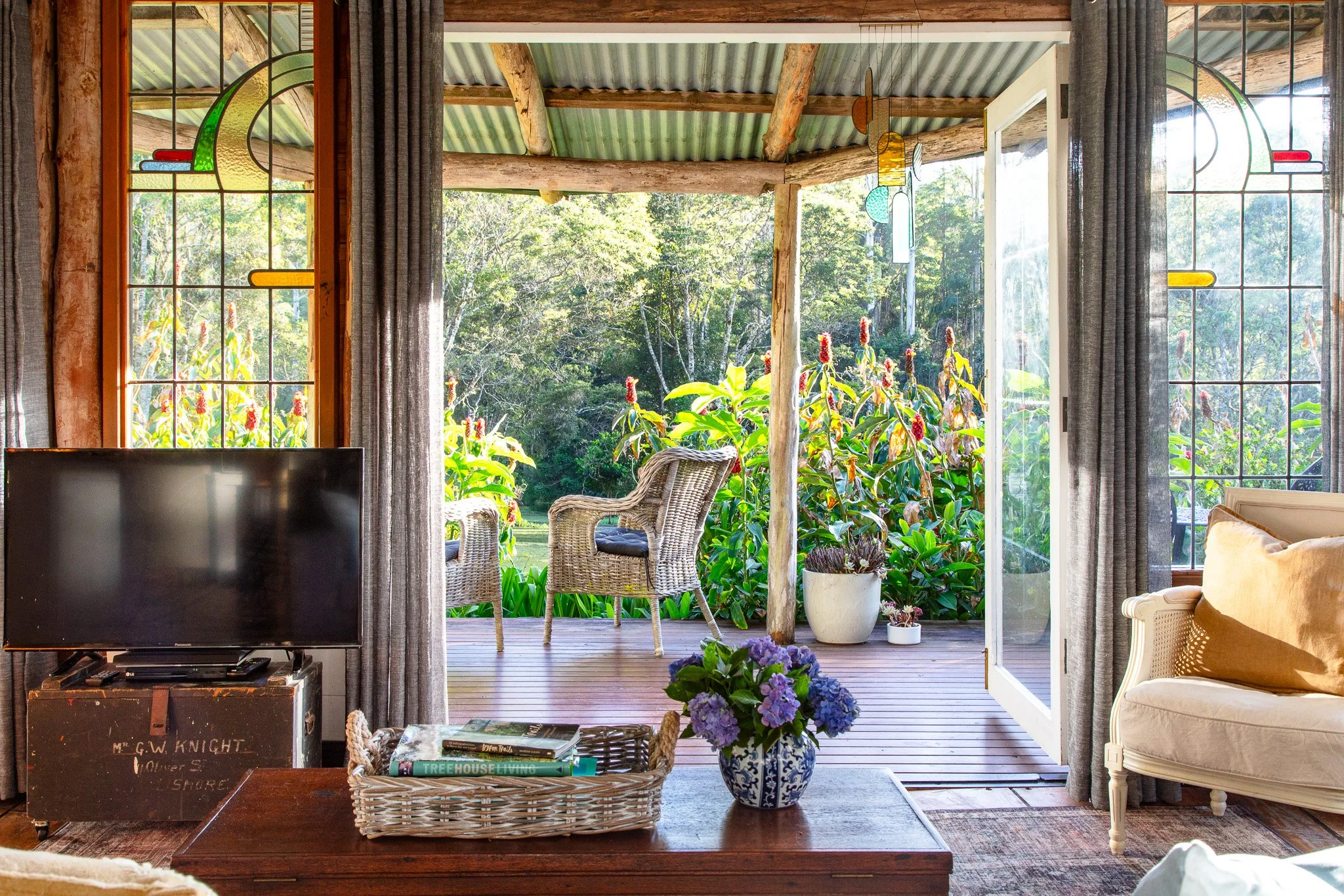 Living room with a flat-screen TV, wicker chairs on a porch, and open sliding glass door leading to a garden with plants and trees.