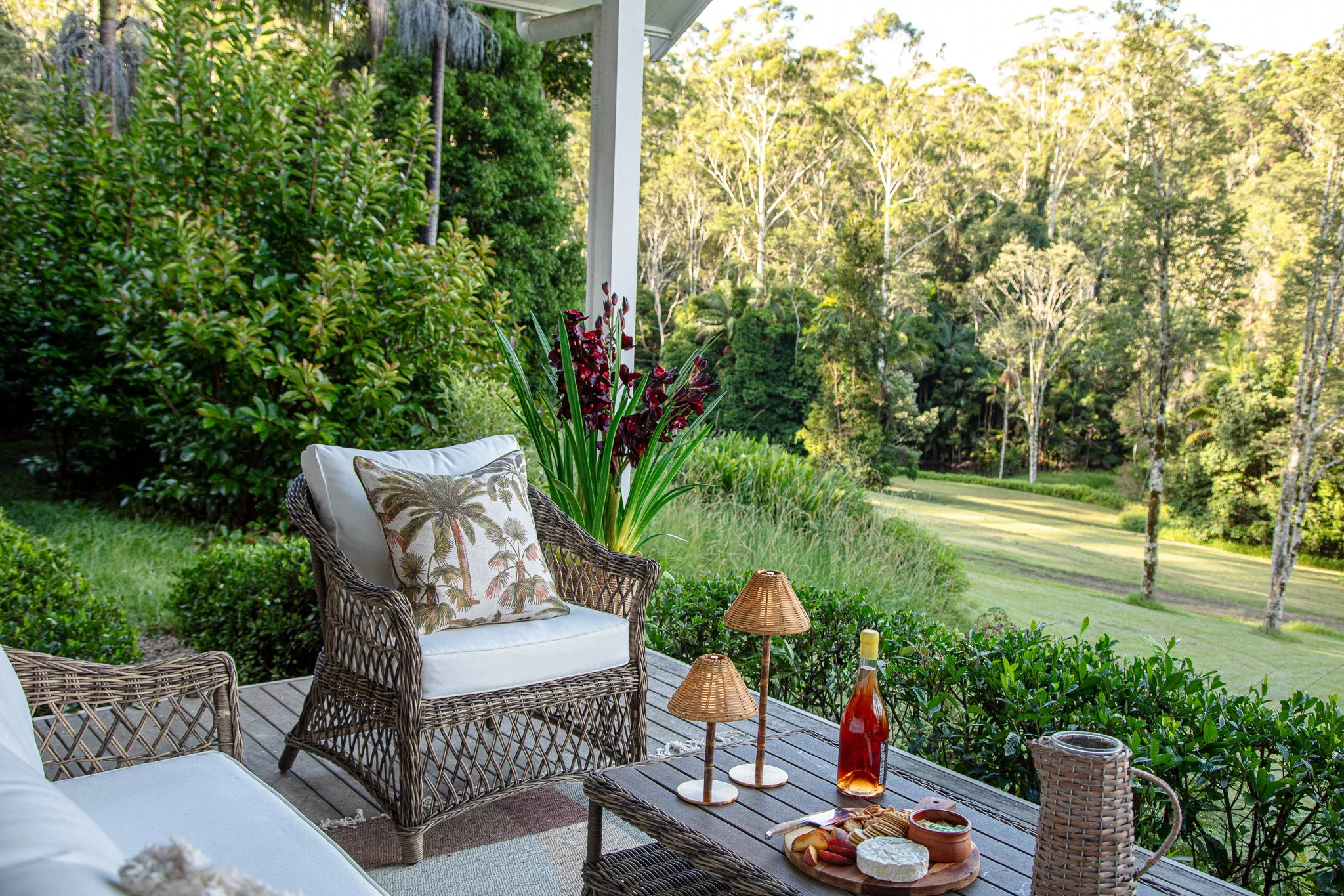 Outdoor patio with wicker furniture, pillows, small lamps, and a bottle of rosé, overlooking a lush green landscape with trees.