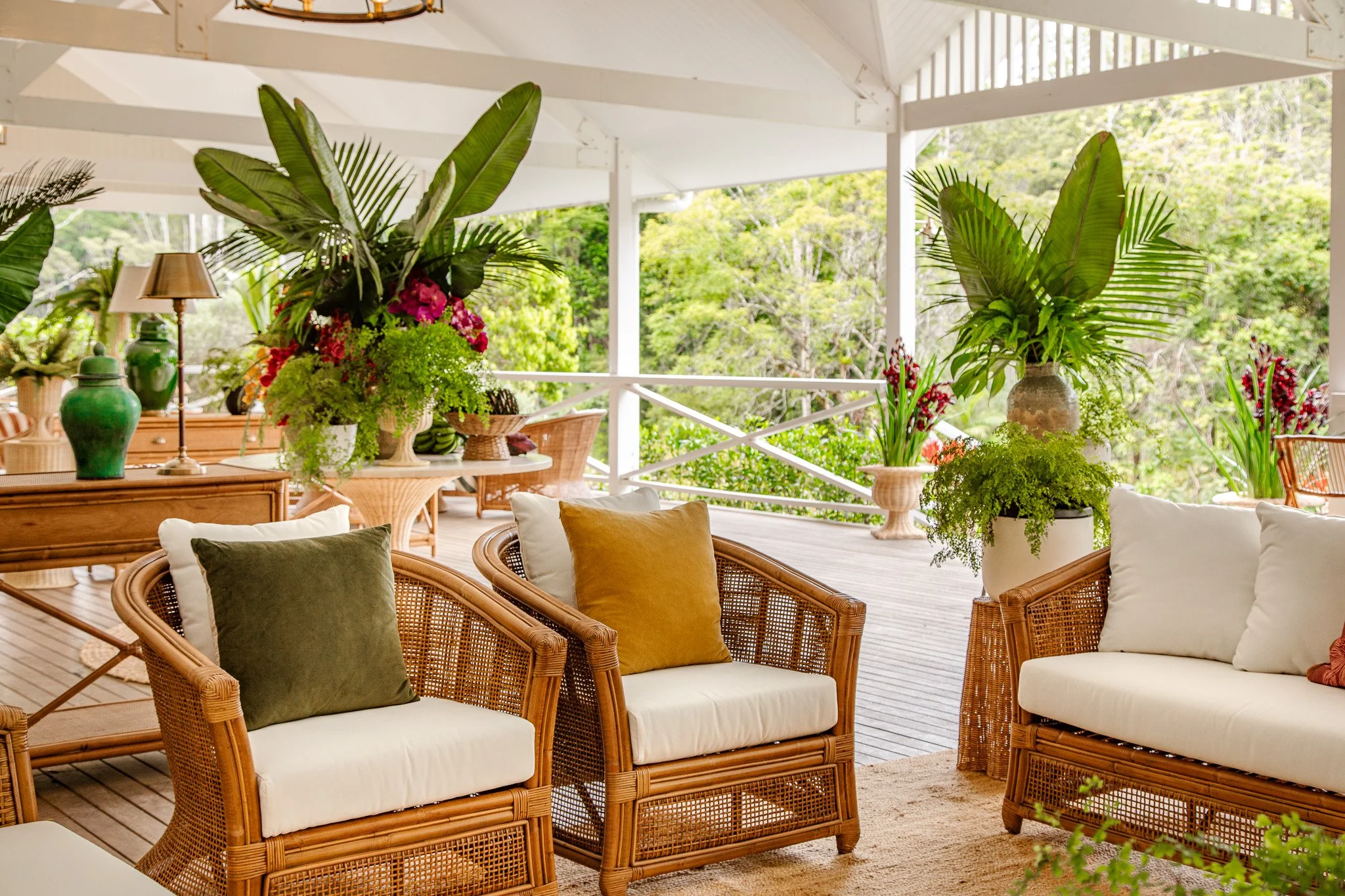 A spacious outdoor patio area with wicker furniture, white cushions, and colorful throw pillows. Large potted plants and floral arrangements decorate the space, with a lush green forest visible in the background.