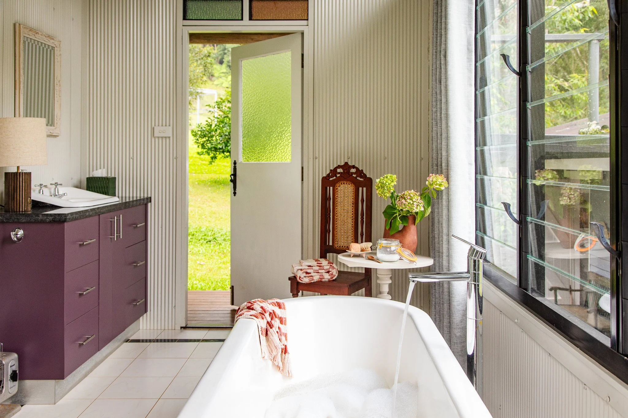 A cozy bathroom with a bathtub filled with bubbles, overlooking a green lawn outside through large window shutters. The bathroom features a purple cabinet with a sink, a wooden chair with a towel, and a small table with flowers and toiletries.