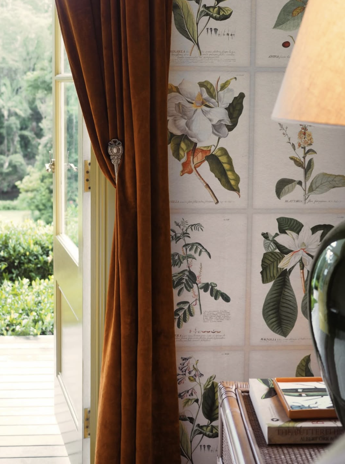 Close-up of a room corner with botanical illustrations on the wall, a partially open door leading outside to a garden, and a side table with a lamp and books.